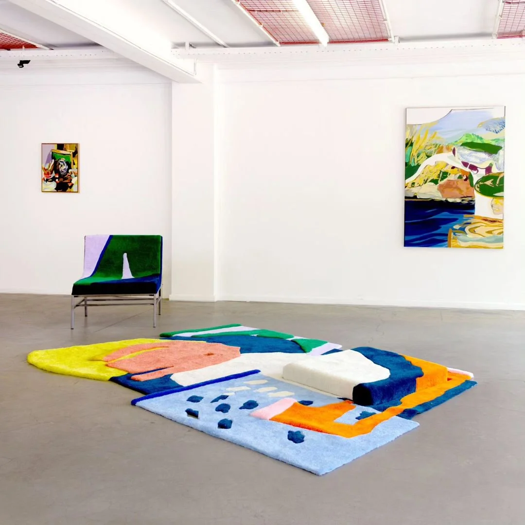Contemporary art gallery featuring colorful, abstract tufted rug on the floor next to a tufted chair and framed paintings on the white walls.