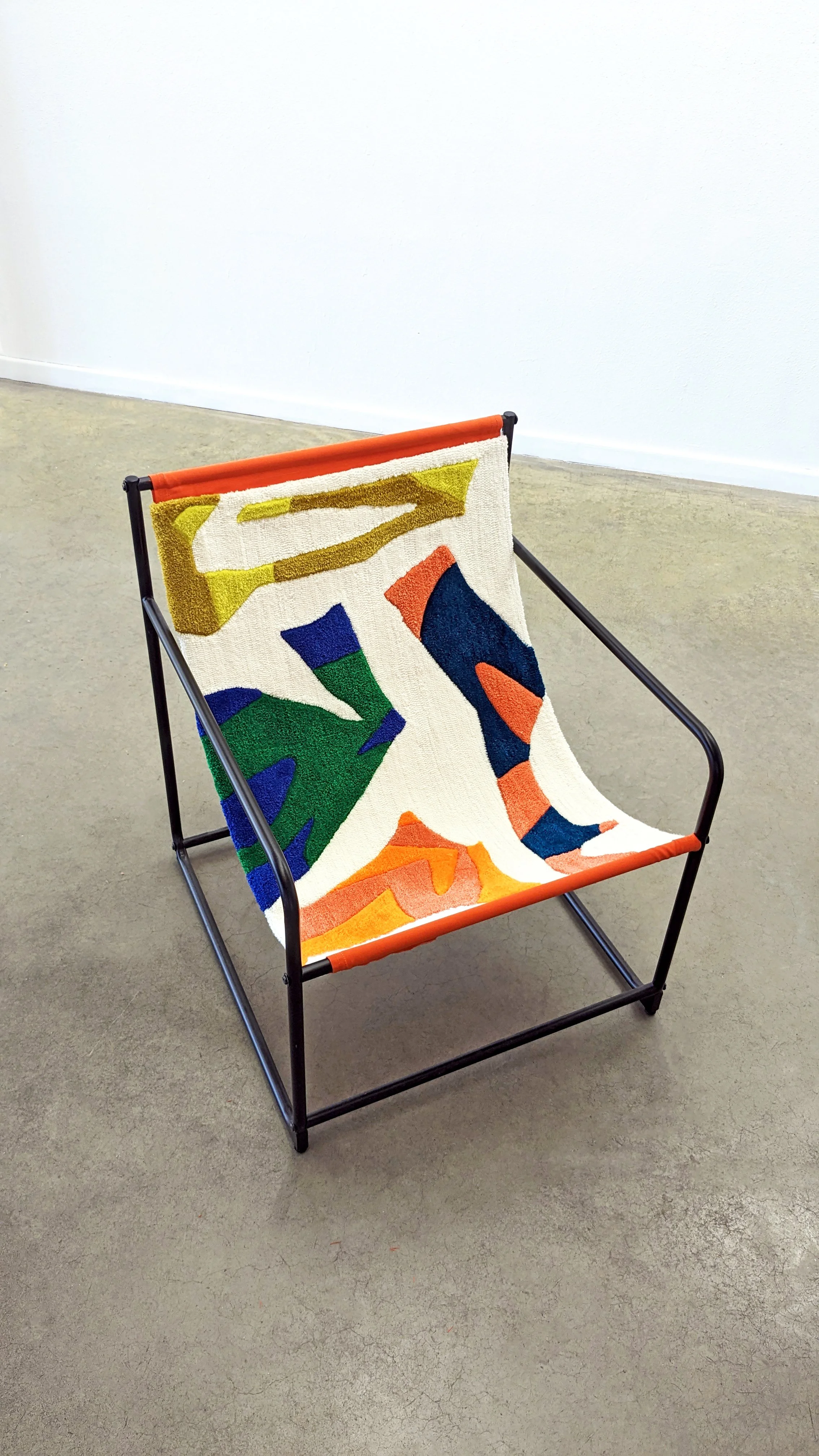 A colorful modern armchair with a fabric tufted  seat featuring abstract geometric patterns in green, blue, orange, yellow, and white, set against a plain light-colored wall and gray floor.