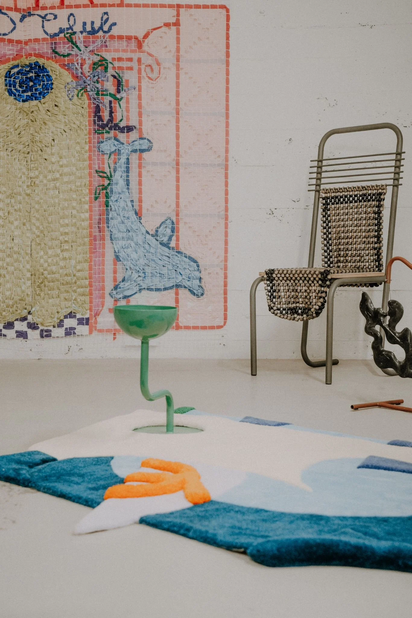 In the foreground, a green table with a winding support holds a green bowl. To the right, a beaded chair stands next to a small, abstract sculpture. Part of a blue and orange tufted rug is visible at the bottom.