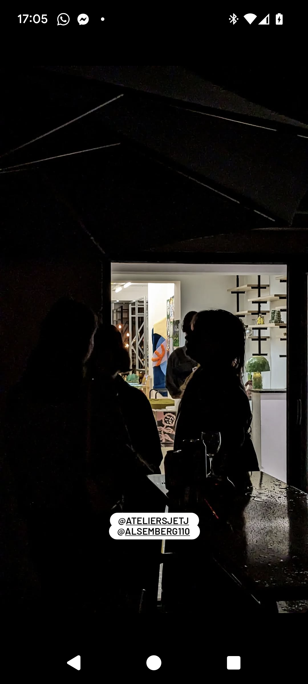 Silhouettes of people inside a dimly lit room, looking through a window into a brightly lit space with shelves and colorful decorations where we can se an tufted art rug on the wall.