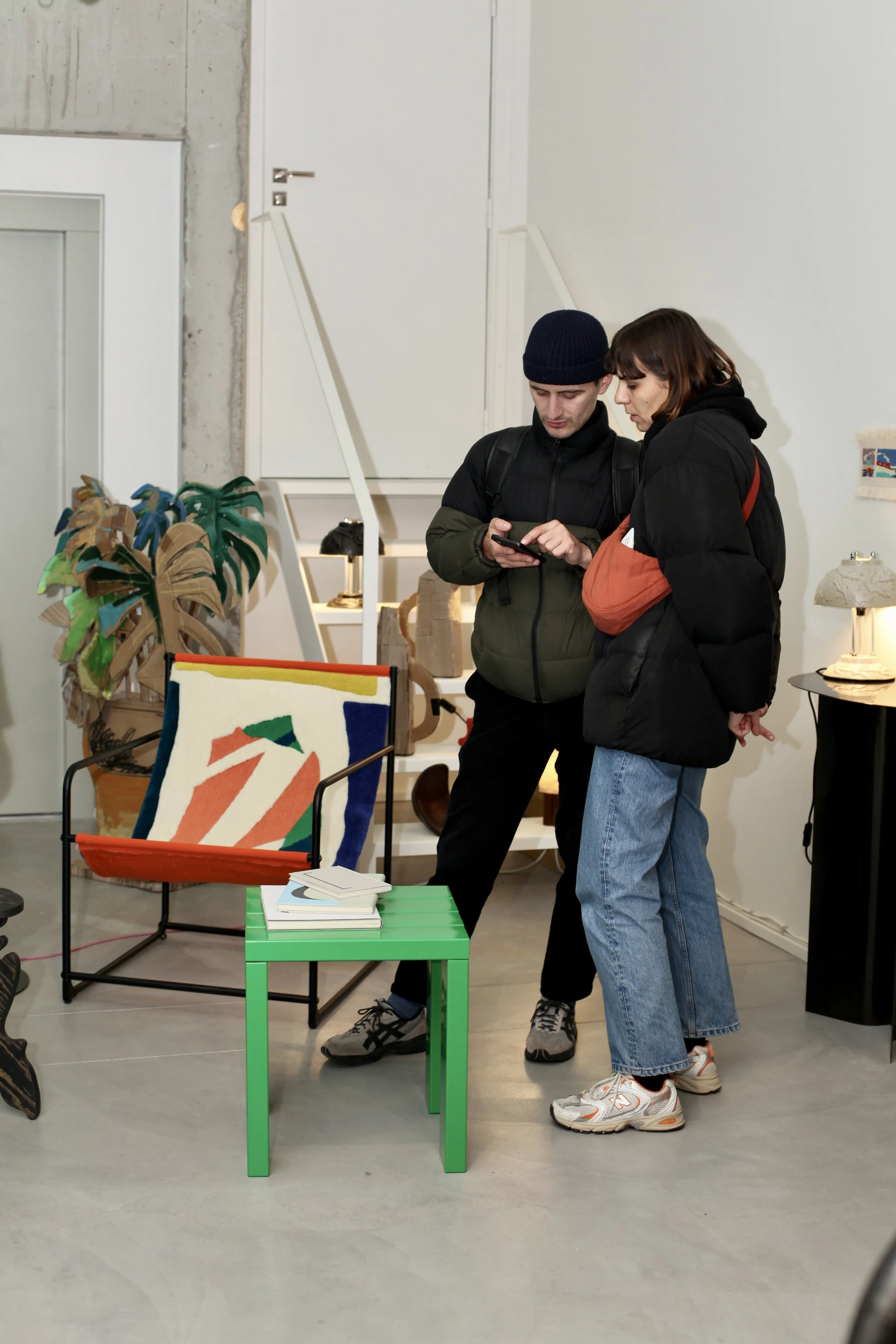 Two young adults, a man and a woman, are standing indoors and looking at a smartphone together. Behind them are some modern furniture and art pieces, including a ceramics and a colorful tufted chair.