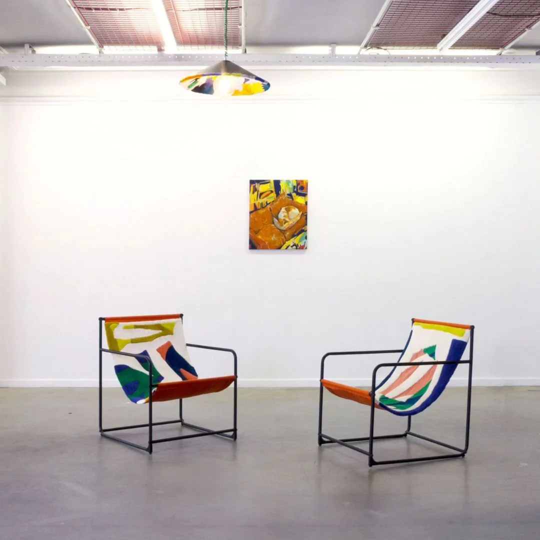 An art gallery with two modern tufted chairs and light suspension with colorful abstract fabric and a small colorful abstract painting on a white wall.