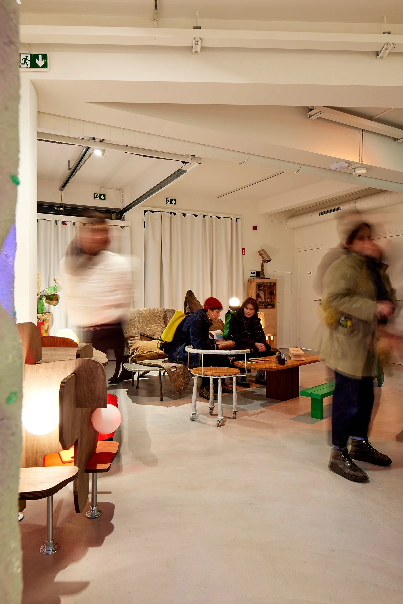 Interior of a cozy living room or cafe with people sitting and talking. There are chairs, a couch, a wooden table, and decorative lighting. Some people are standing and blurred due to motion.
