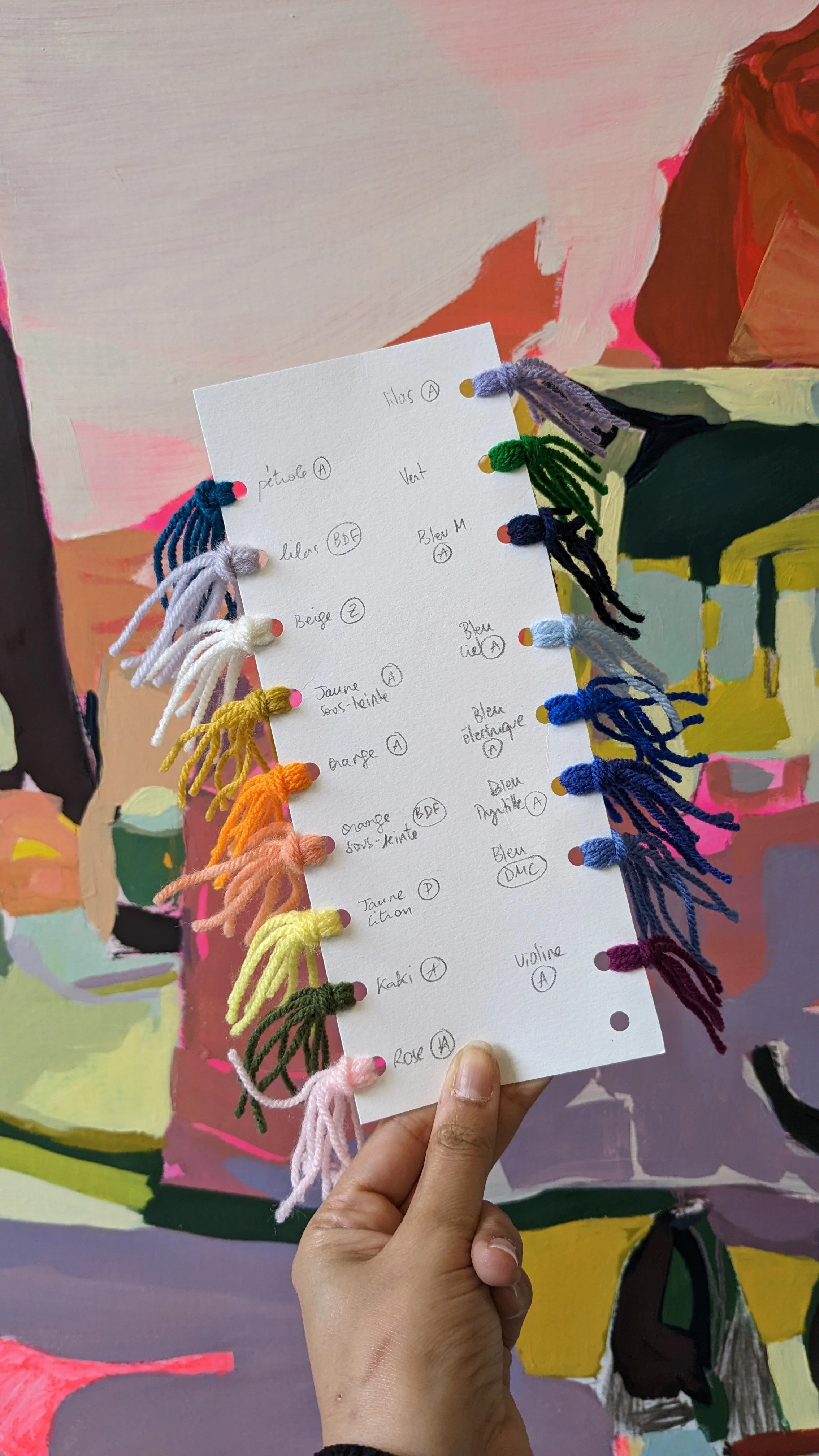 A hand holding a color wool thread sample card with handwritten notes, in front of a colorful painted background.