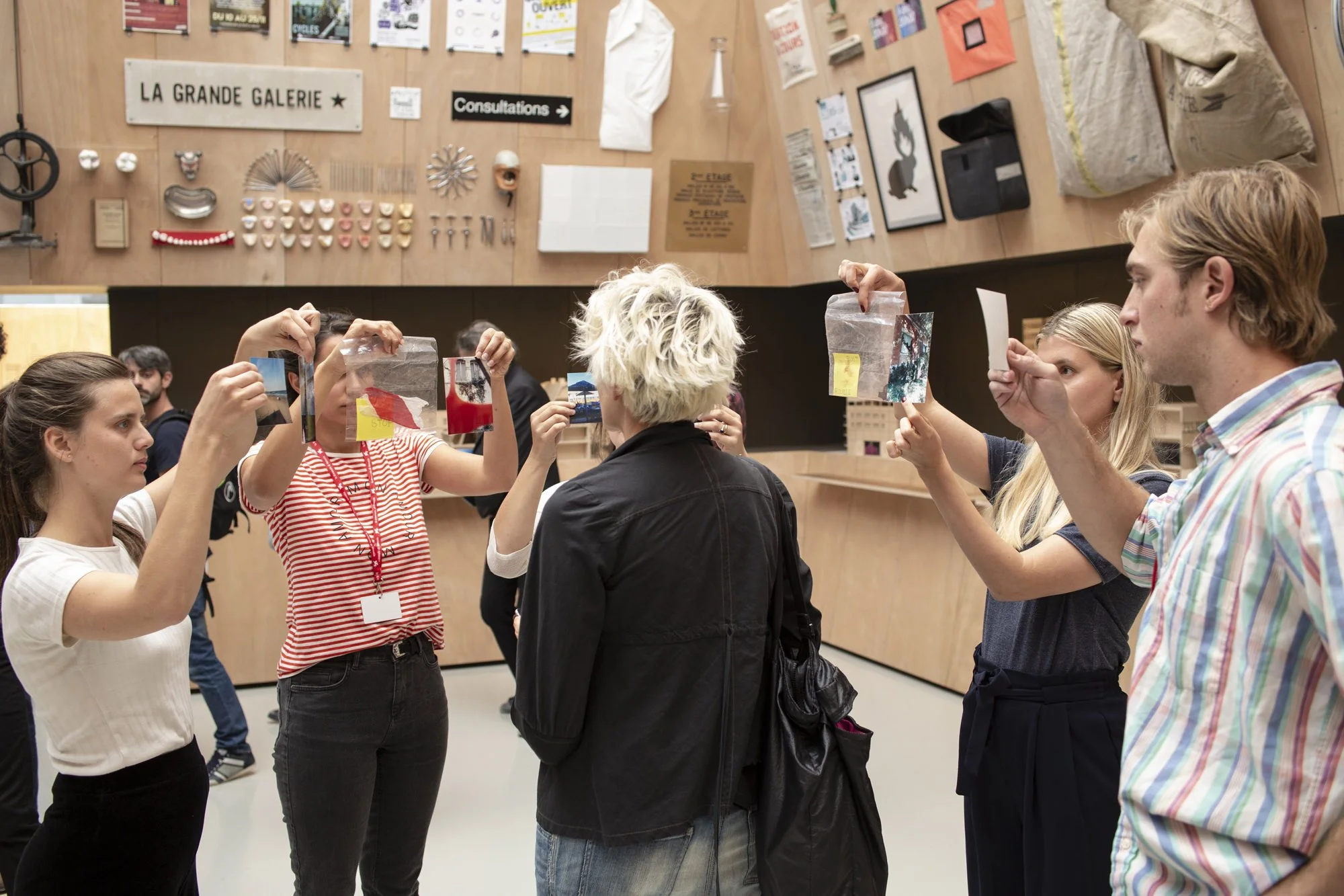 A group of people participating in an art gallery event, standing in front of a wall with artwork and signs. They are taking photos or looking at postcards featuring the artwork.