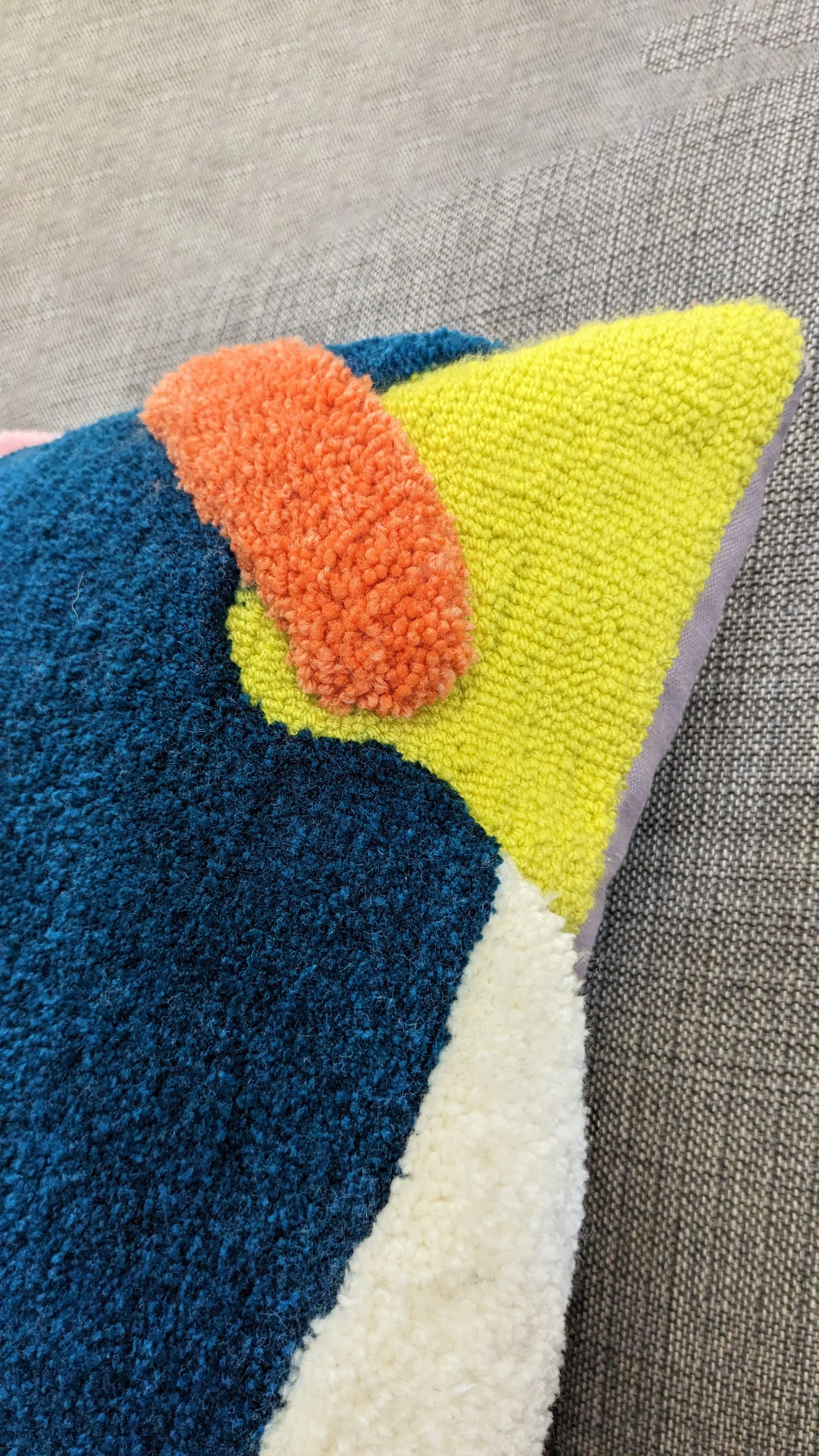Close-up of a colorful, textured tufted cushion with various colors including orange, blue, yellow, white, and gray, placed on a gray fabric surface.