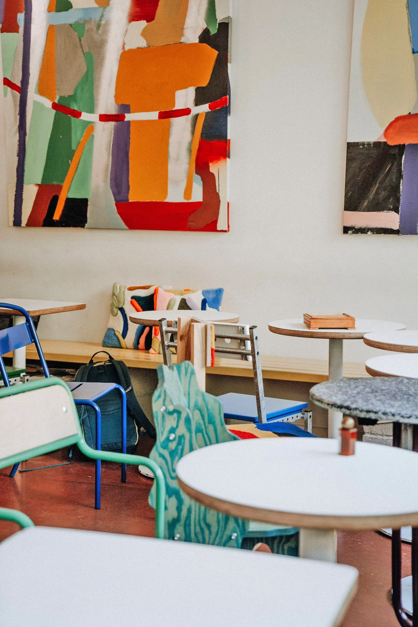A restaurant with colorful chairs, tables, tufted pillows, and abstract paintings on the walls.