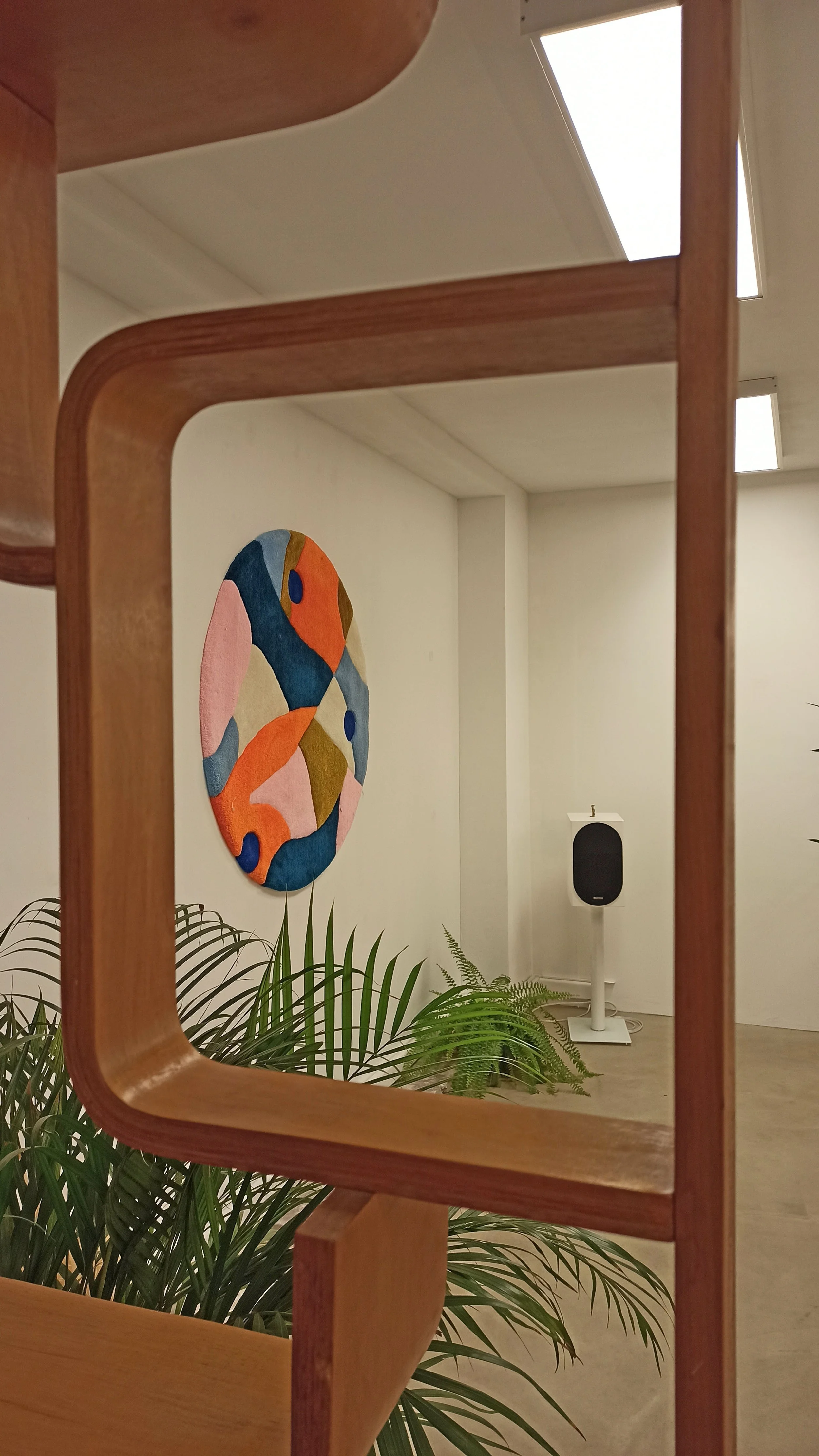 An art gallery room with a colorful abstract tufted rug tapestry on the wall, a black speaker on a stand, and indoor plants in the foreground, viewed through the wooden frame of a chair.