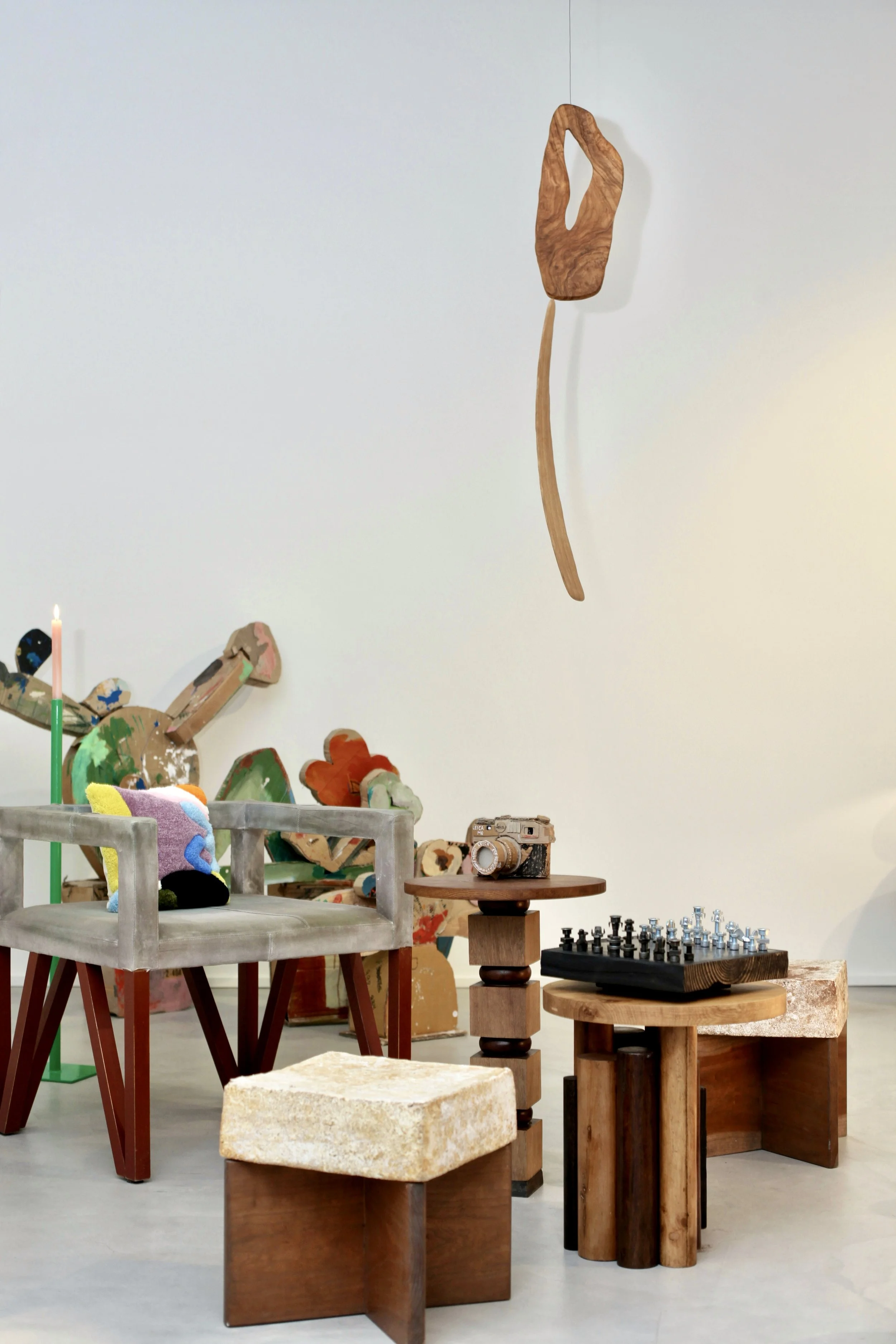 Various handcrafted furniture pieces and decorative items in an art gallery or store, including a plush armchair, wooden tables, and a wall art installation resembling a stylized shape, with a chessboard on one table.