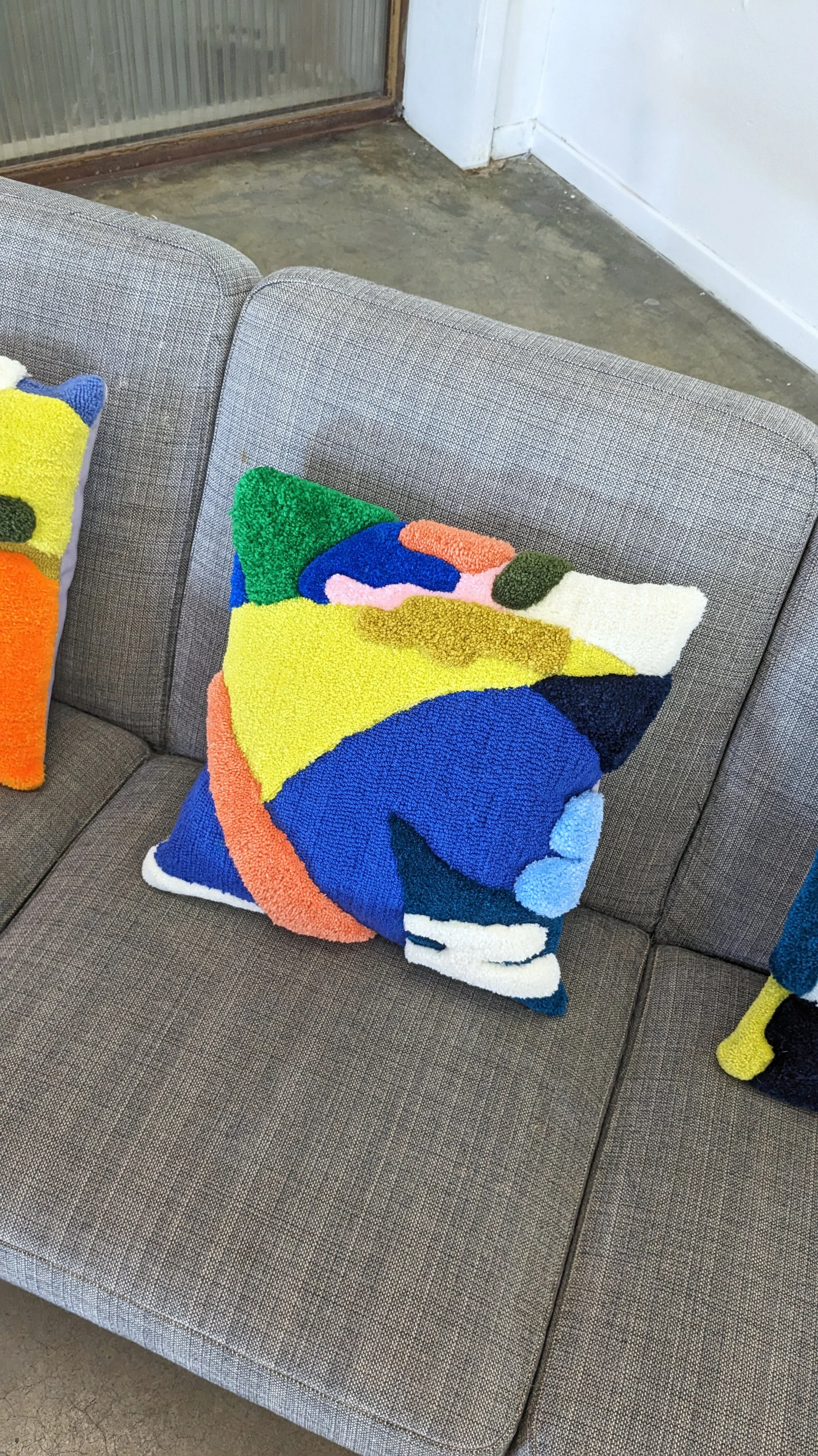 Colorful abstract tufted throw pillows on a gray fabric sofa, next to a concrete floor and a white wall.