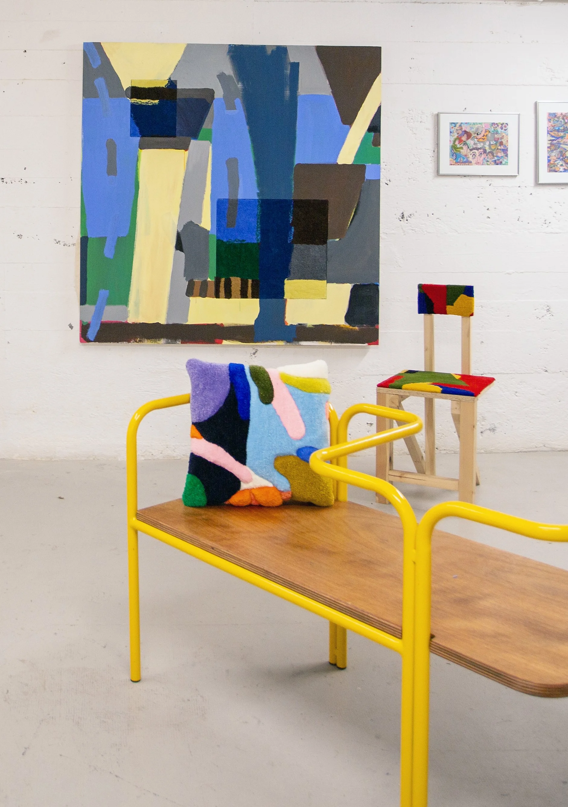 An art gallery showcases an abstract painting with bold colors and geometric shapes on a white wall, accompanied by a wooden chair with colorful fabric and a yellow metal frame, as well as a second chair with a colorful cushion. Two smaller framed ar