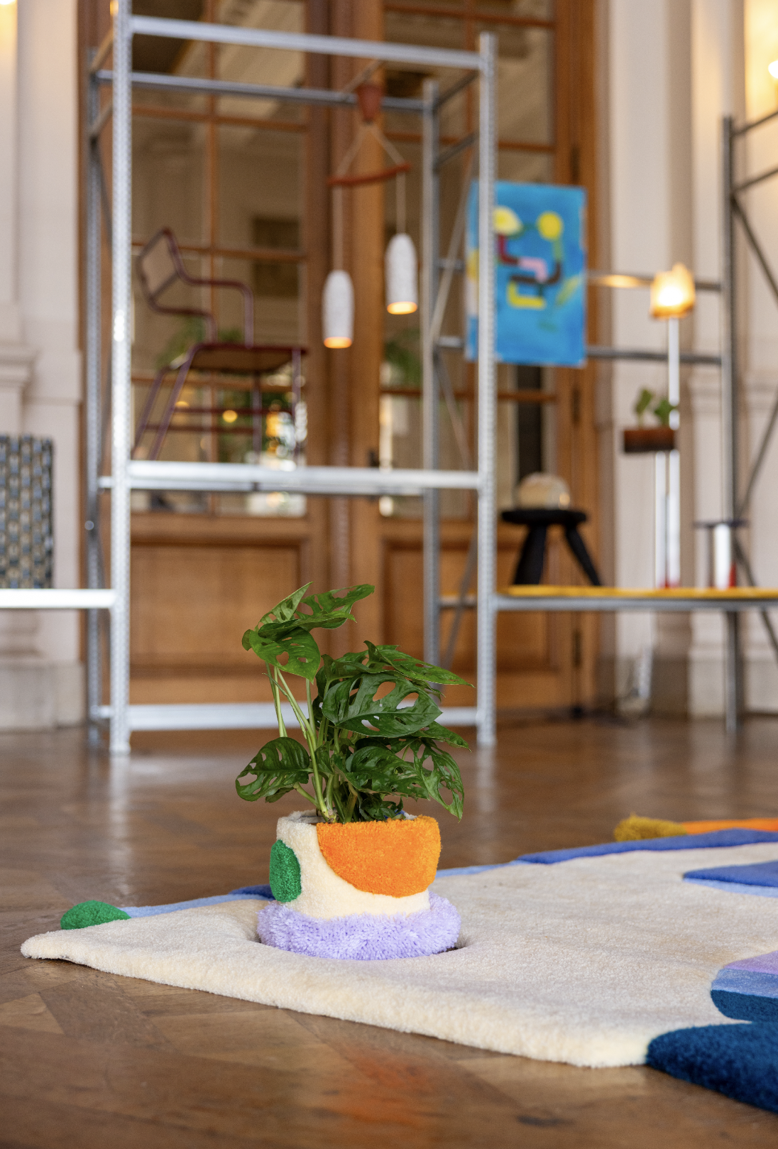 A potted indoor plant with large green leaves in a colorful, textured tufted plant cover with two pile heights on a patterned tufted rug, with various modern furnishings and decor in the background.