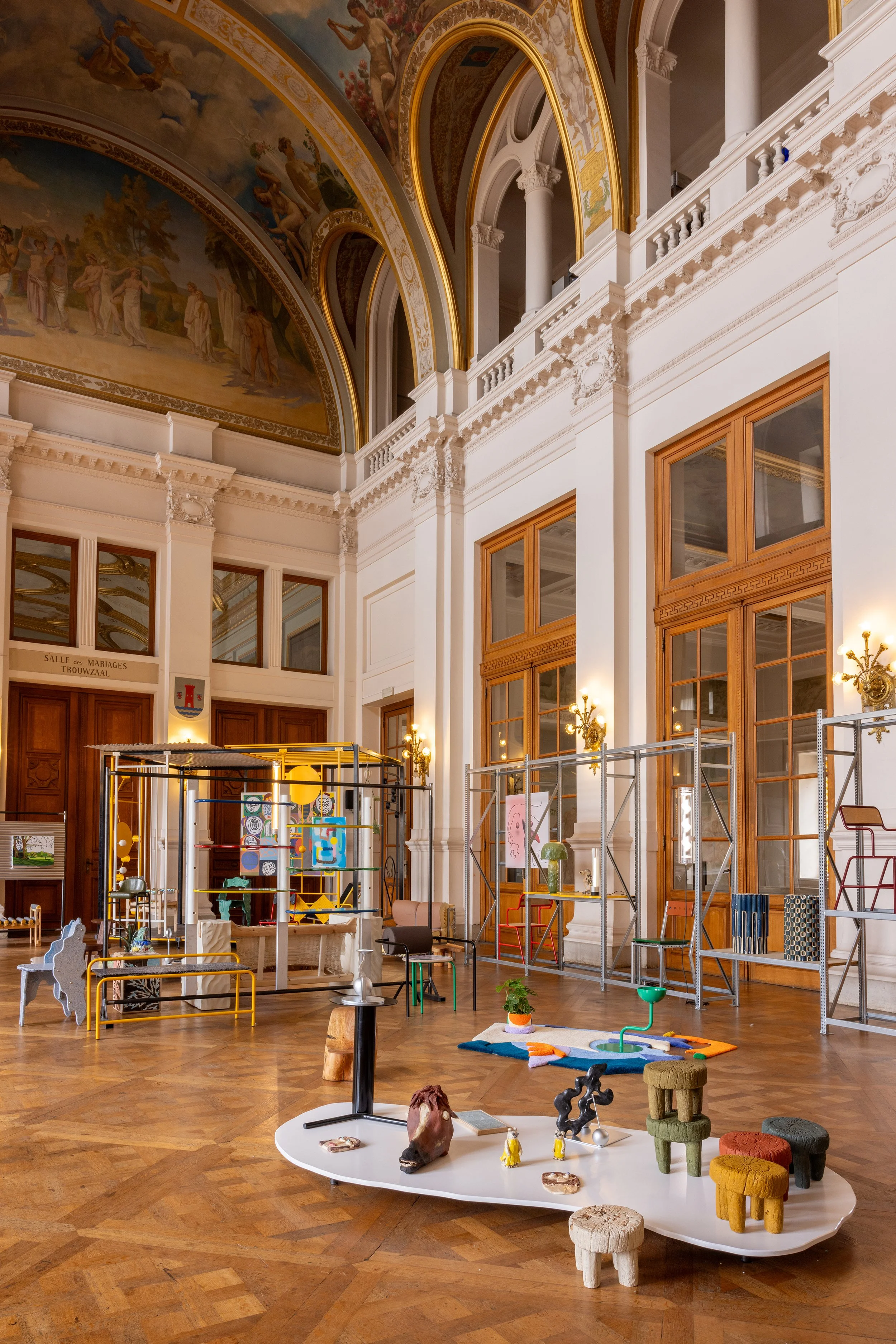 Interior of an elegant, historic building with ornate ceiling murals, tall windows with wooden frames, and display of modern art and sculptures on scaffolding and stands, with hardwood floors and vintage lighting fixtures.