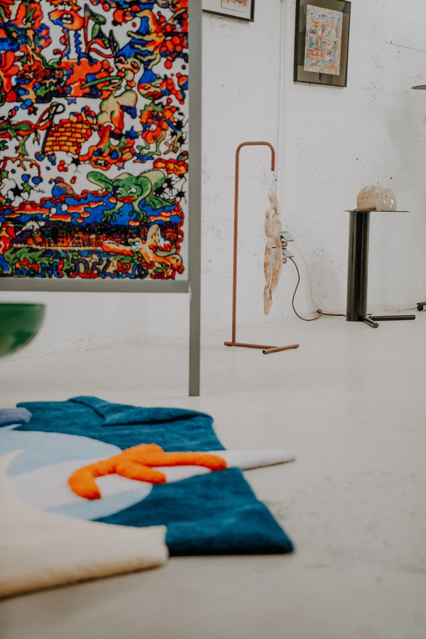 Interior of an art gallery with colorful artwork on the wall, a sculpture on a stand, and a tufted rug with blue and orange wool.