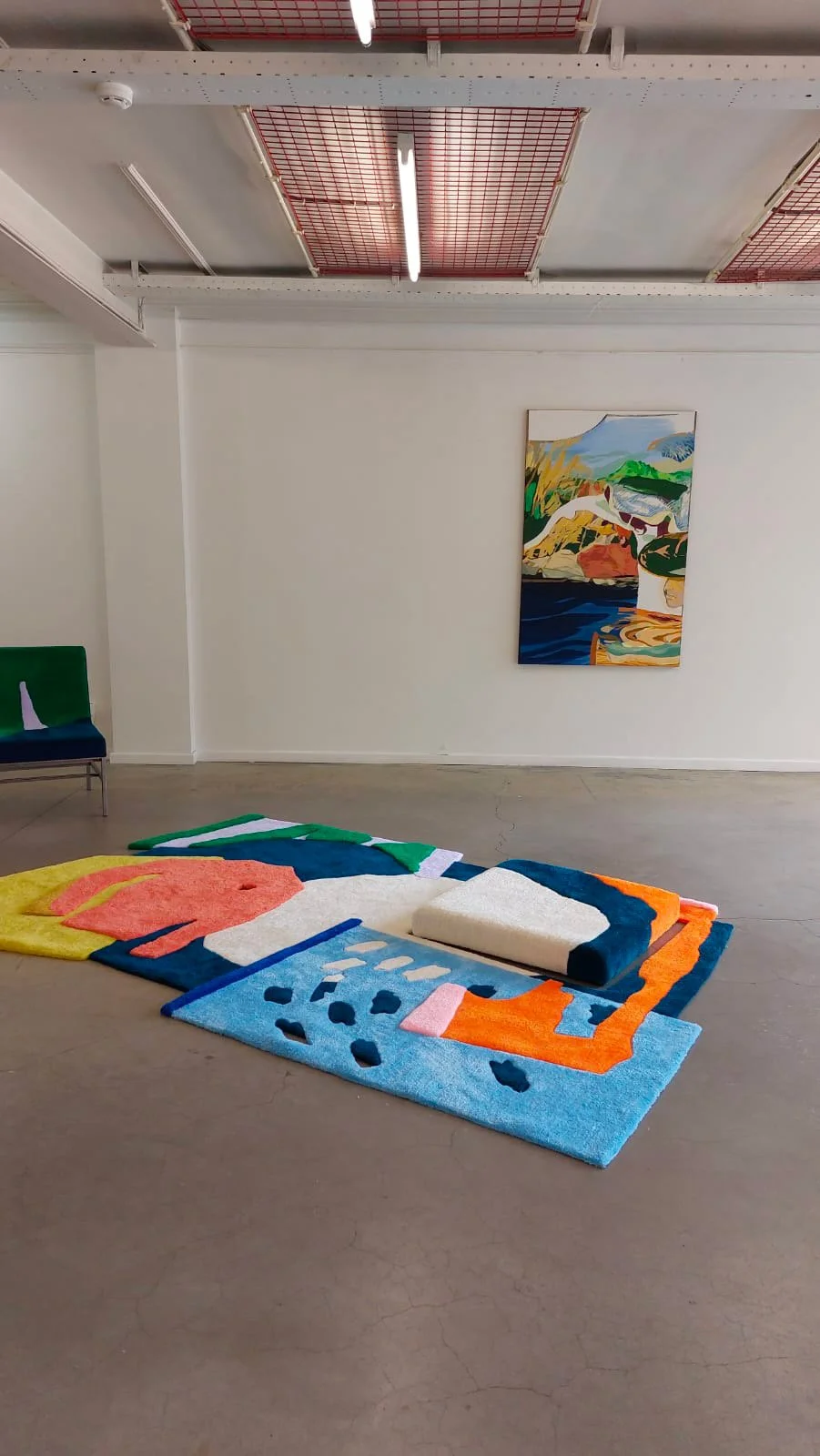 An art installation featuring a colorful tufted rug arranged on the floor resembling a face, with a white cushion serving as the nose. A colorful landscape painting hangs on the wall behind the installation.