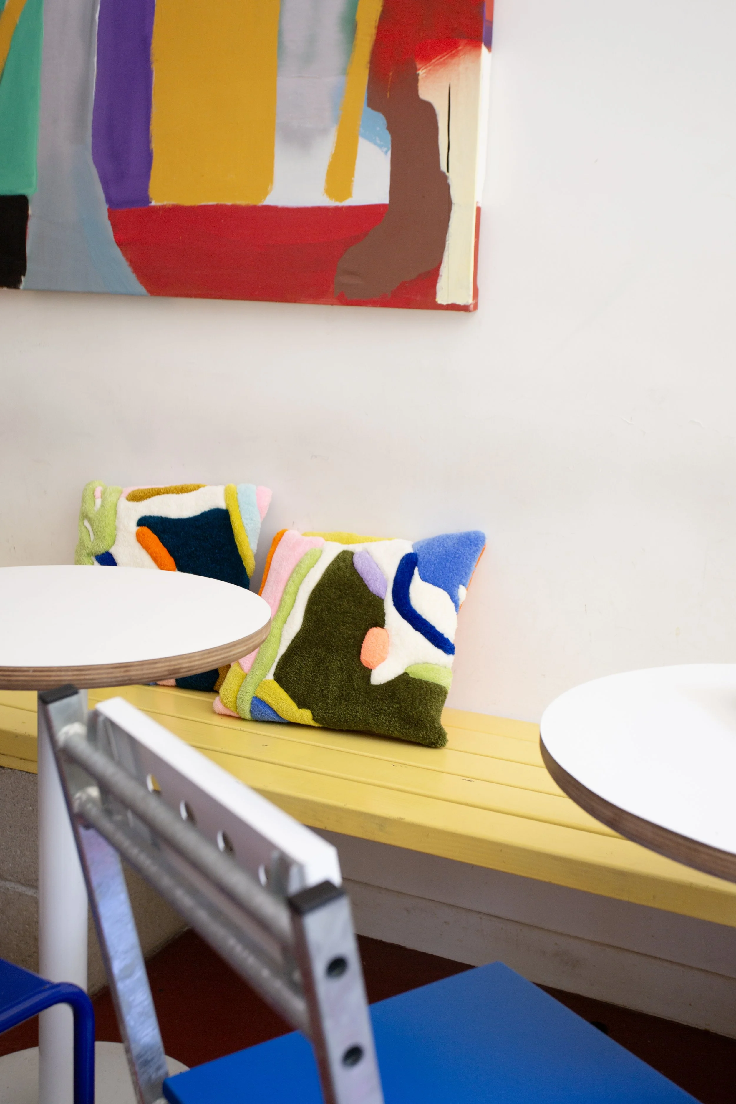 Colorful abstract painting hangs on a white wall above a yellow bench. Multiple patterned tufted cushions / pillows with abstract designs are on the bench, and part of a round white table and metal chair are visible in front.