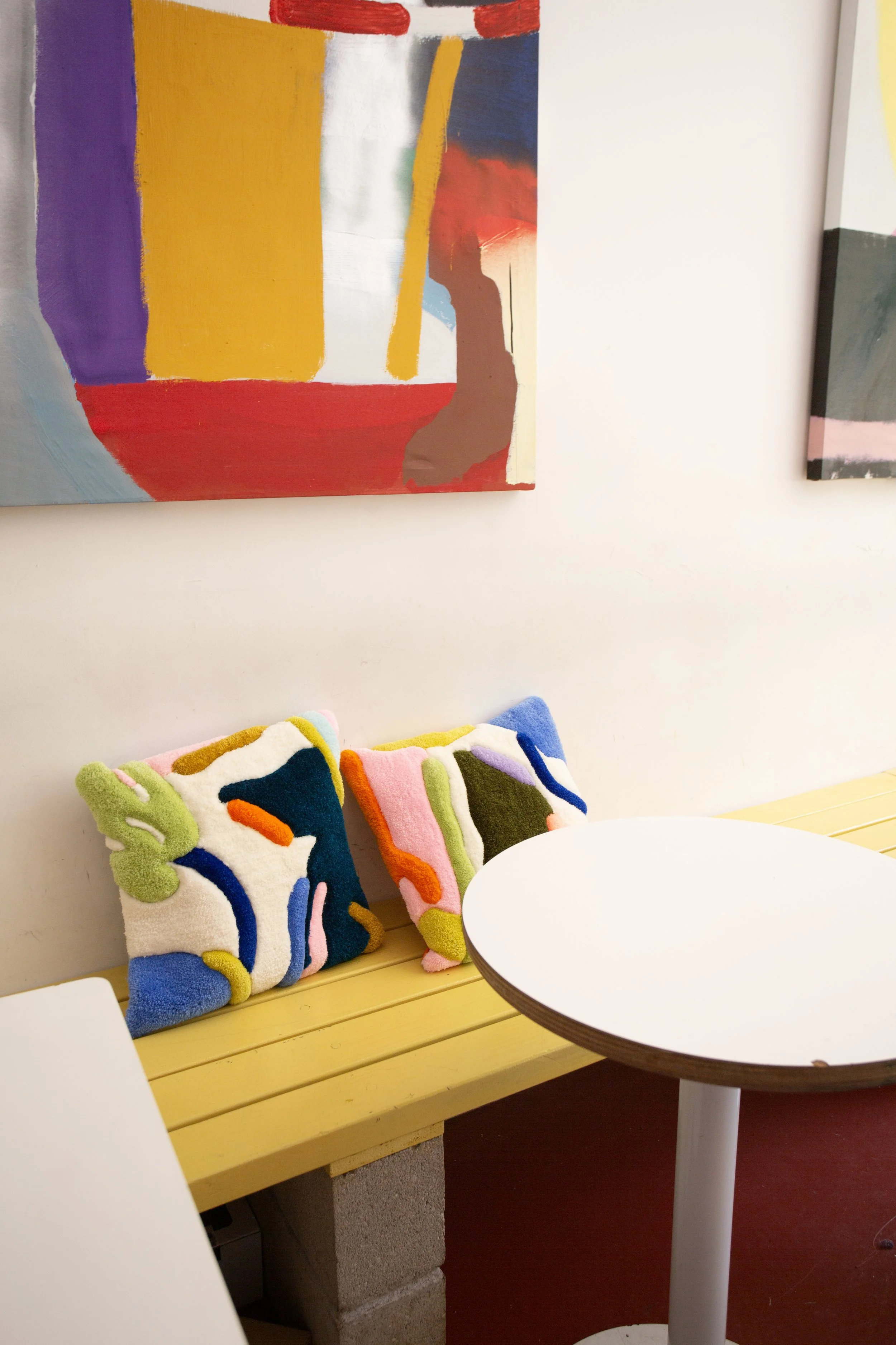 A display of three colorful abstract-patterned tufted pillows on a yellow wooden bench in a room with a white wall and framed artwork. A white round table is in the foreground.