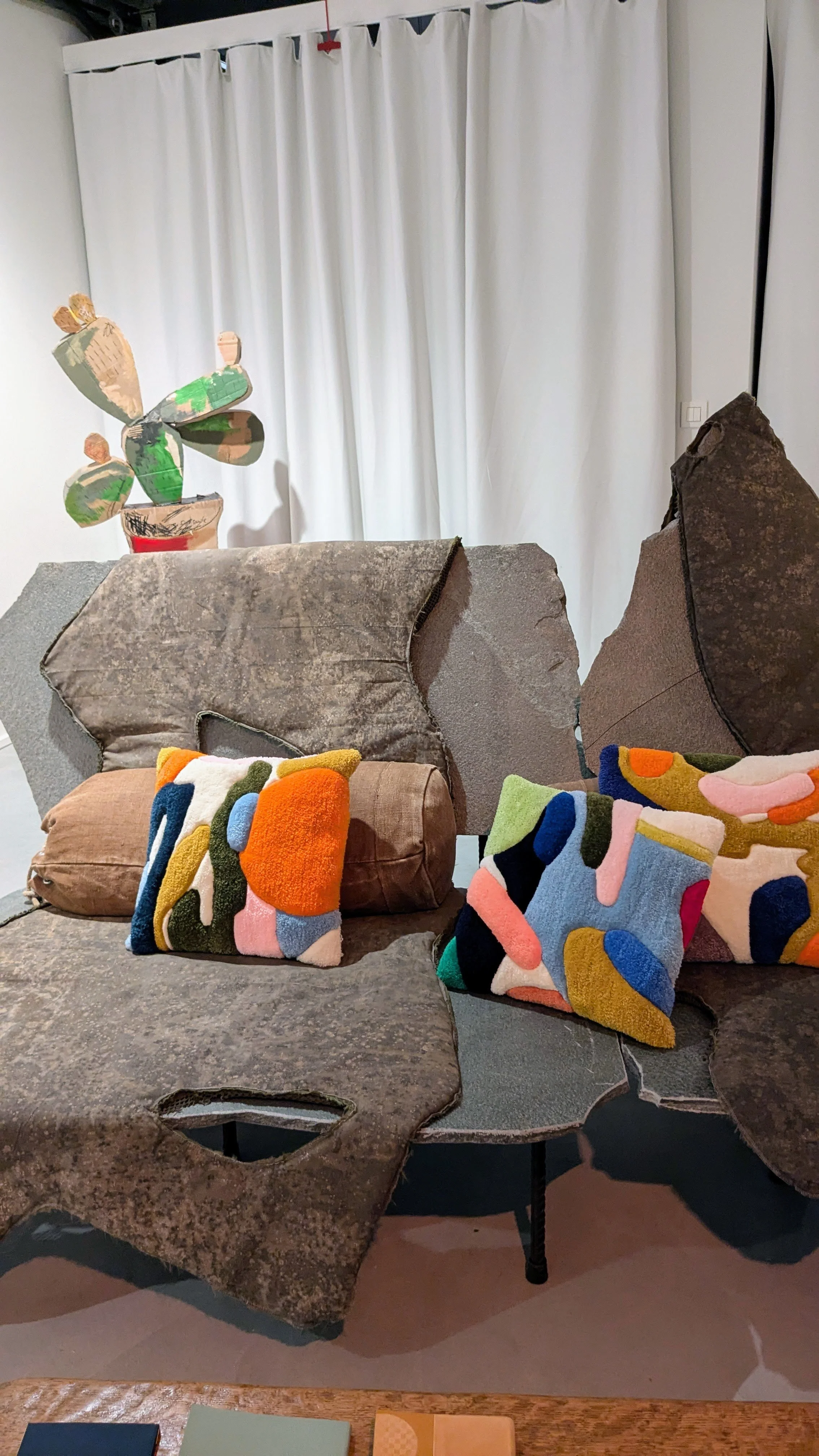 Living room scene in exhibition featuring a sofa with colorful abstract tufted patterned cushions, overgrown tufted pillows, a cardboard cactus decoration on a stand, and a plain white curtain in the background.