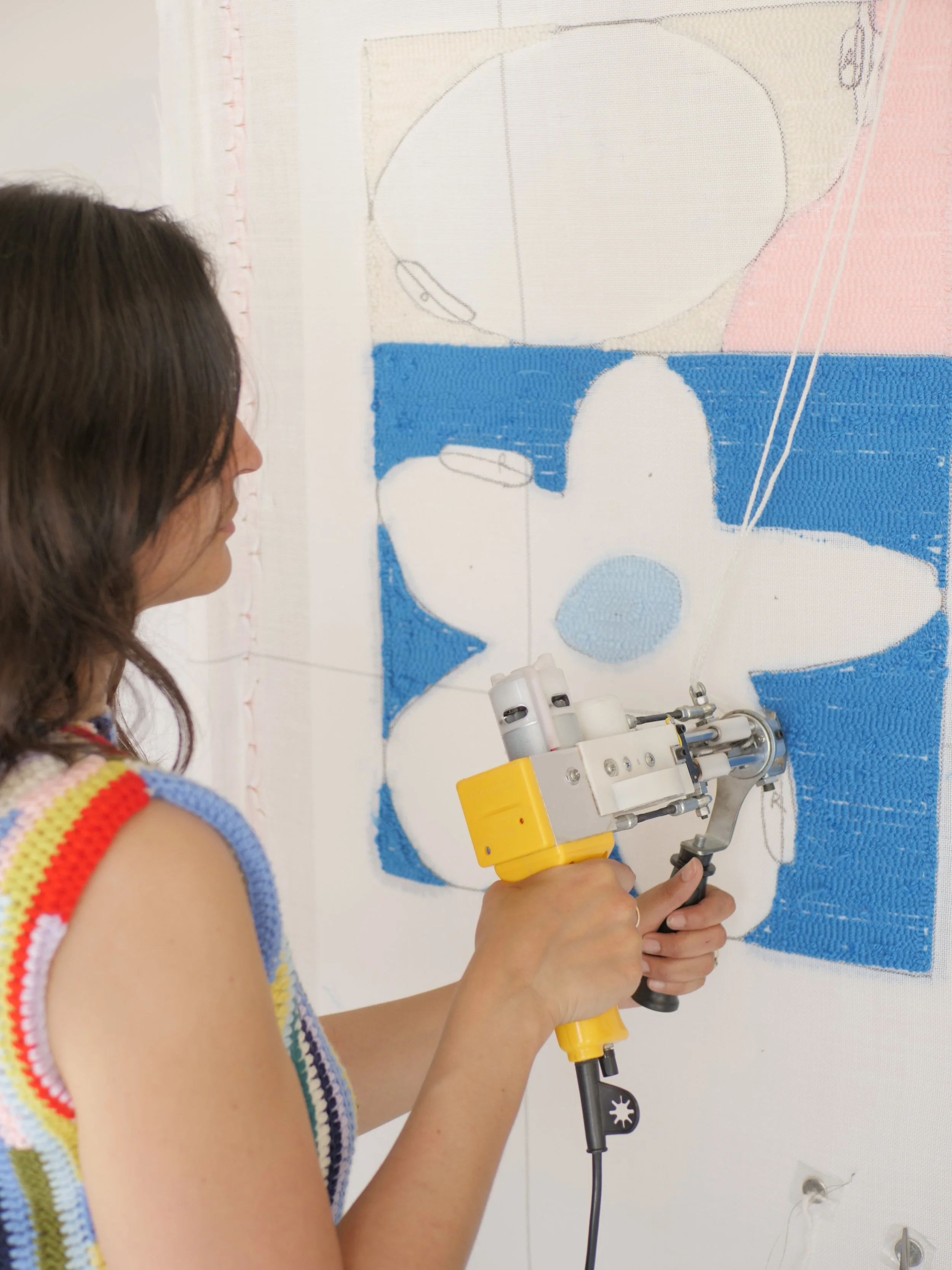 A person holding a yellow and black tufting gun while working on a tufting frame. The cloth has a sketch of an abstract flower illustration and is partially tufted with blue and pink wool.