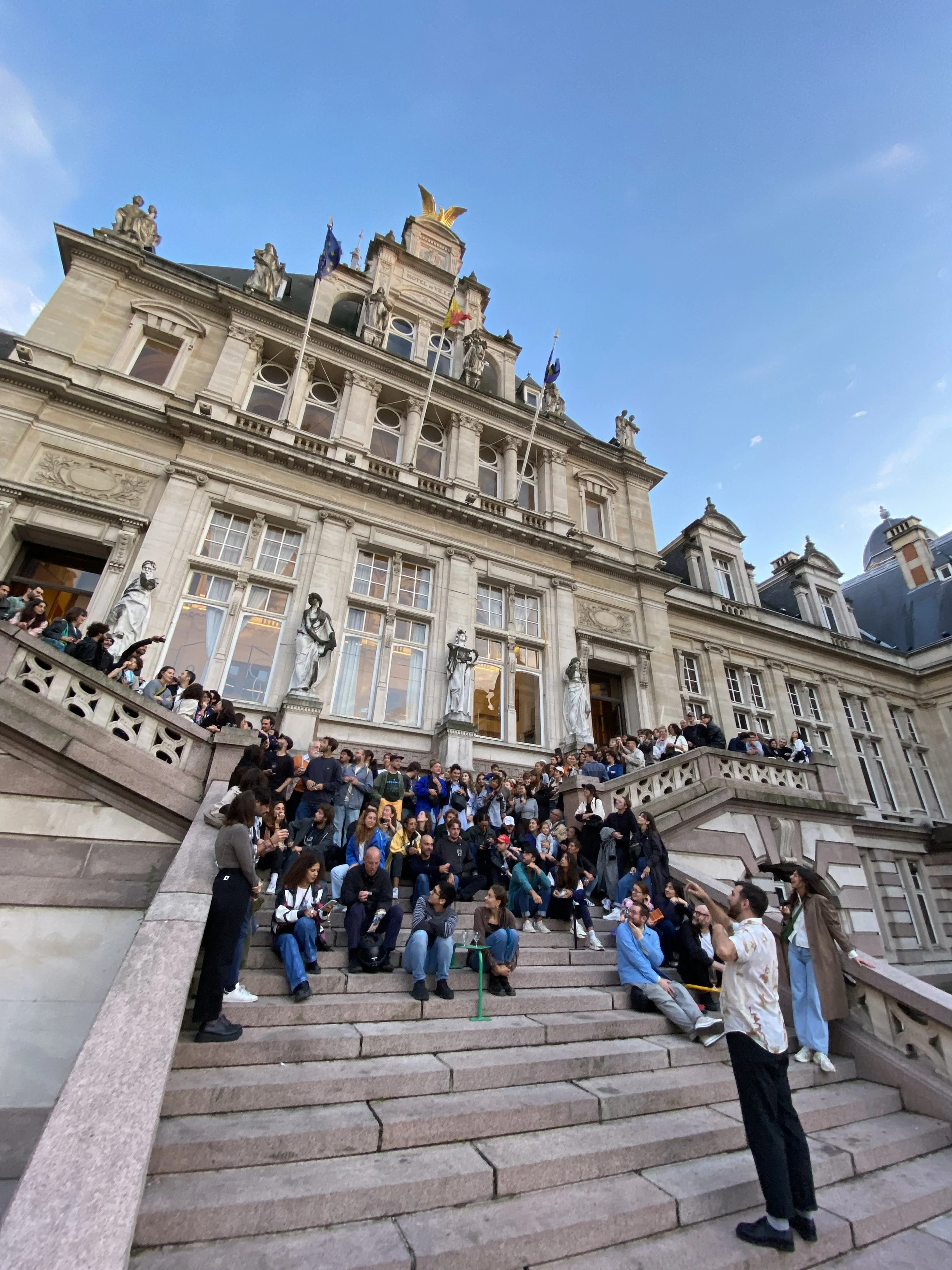 People sitting and standing on a large stone staircase outside the historic building "La maison communale de saint-gilles" with statues and flags, under a clear blue sky.
