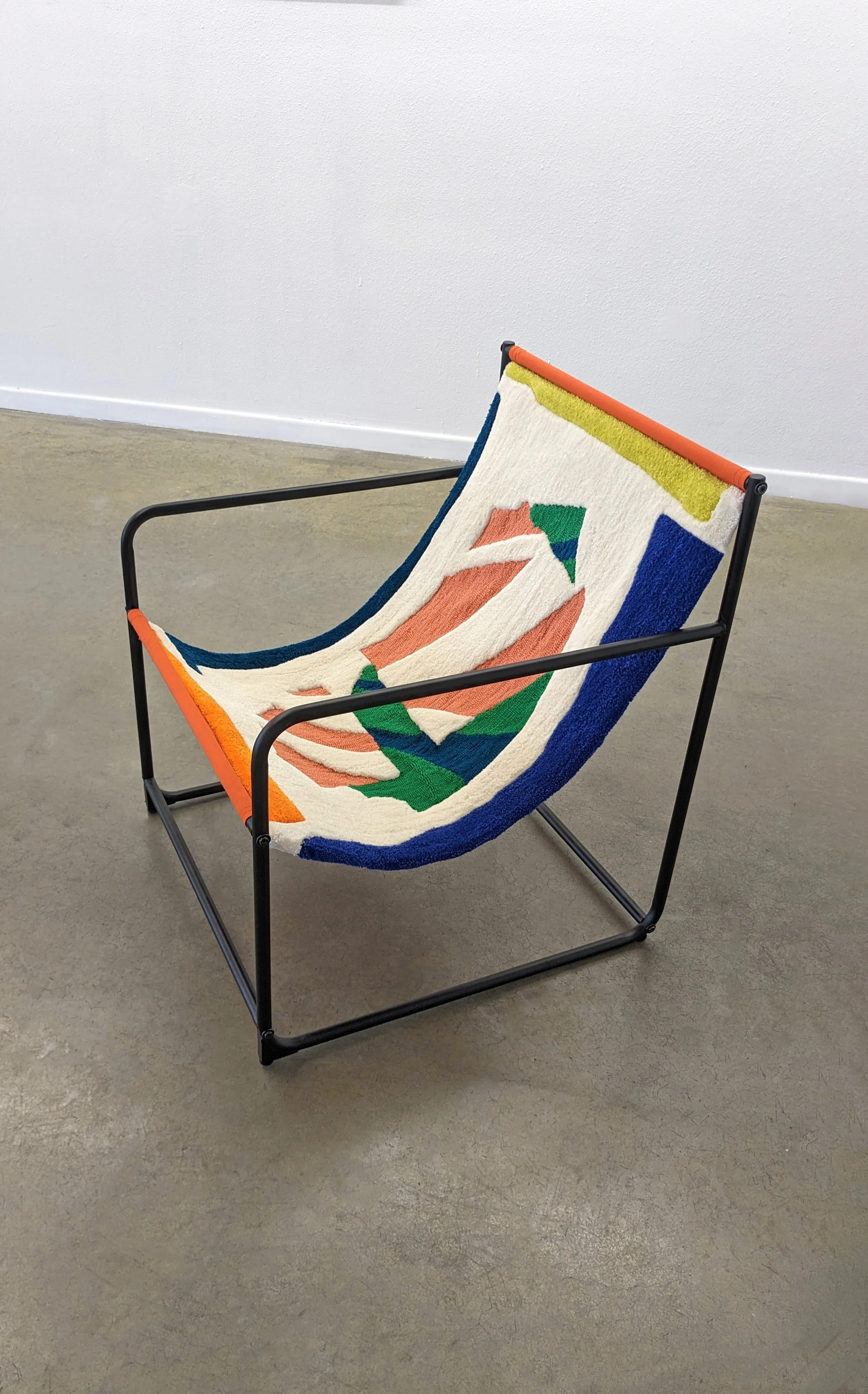 A chair with a colorful, abstract textile seat tufted and backrest featuring green, blue, orange, and white colors, situated against a plain white wall and sitting on a gray concrete floor.