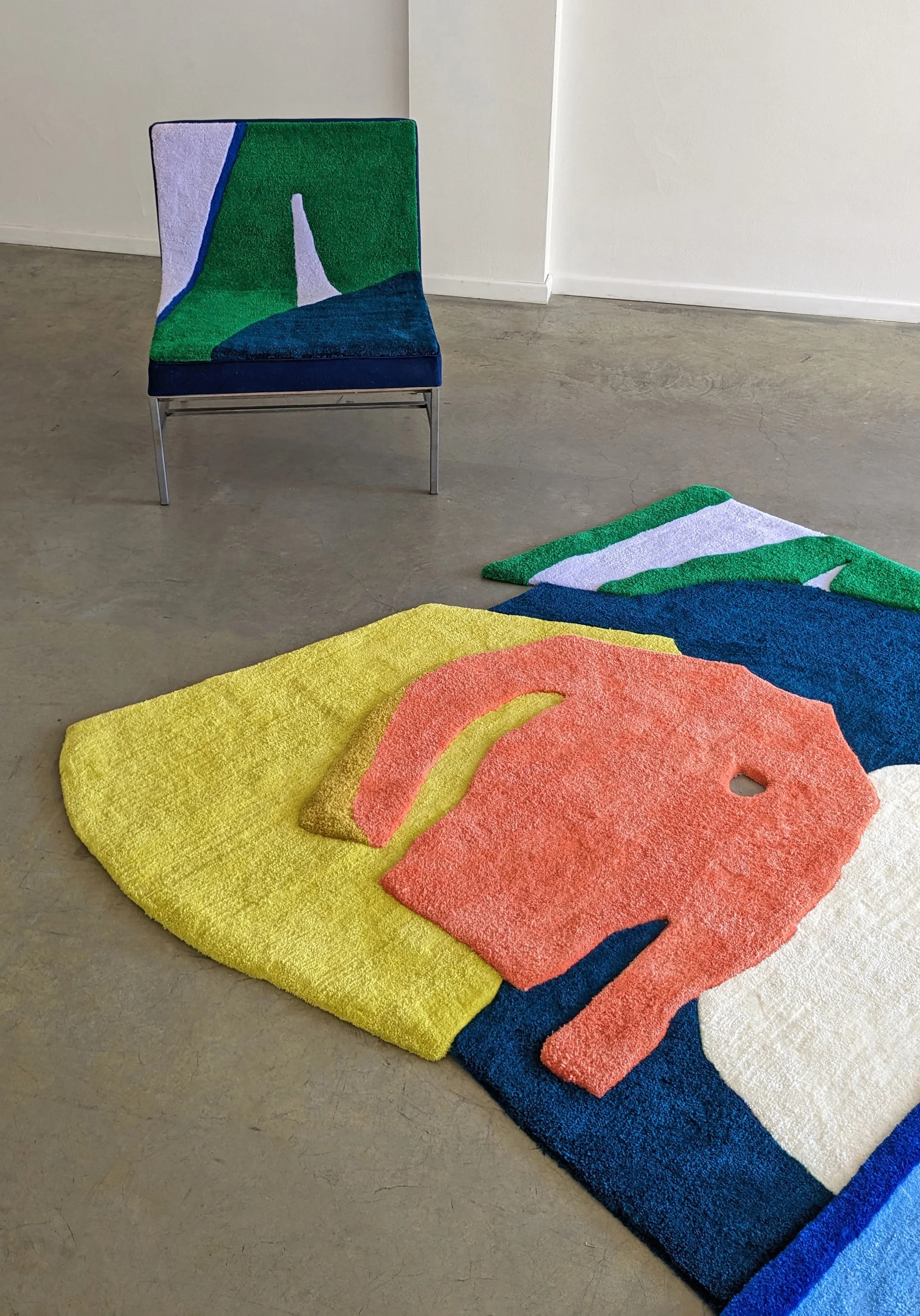 Colorful tufted rug with abstract design in colors like yellow, orange, pink, blue, green, white, and black, laid out on a concrete floor beside a chair with similar design and colors.