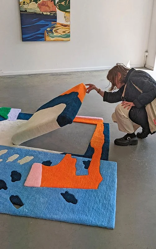 Person bending over to view a colorful, abstract rug in an art gallery.