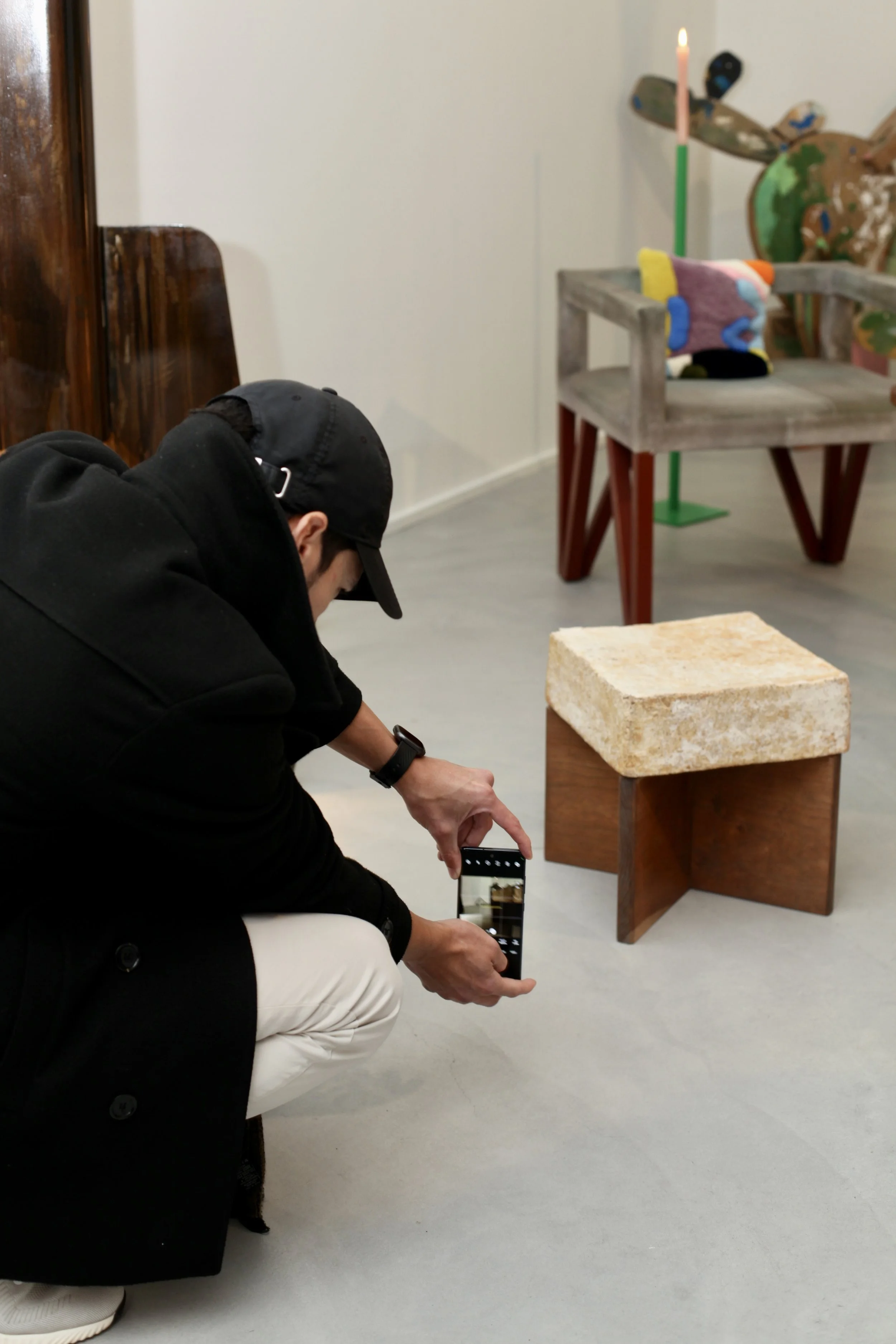A person wearing a black jacket and a black cap is kneeling and taking a photo with a smartphone of a middle-sized art piece on a wooden pedestal. The art piece appears to be a block made from a textured, stone-like material. Behind the artwork, ther