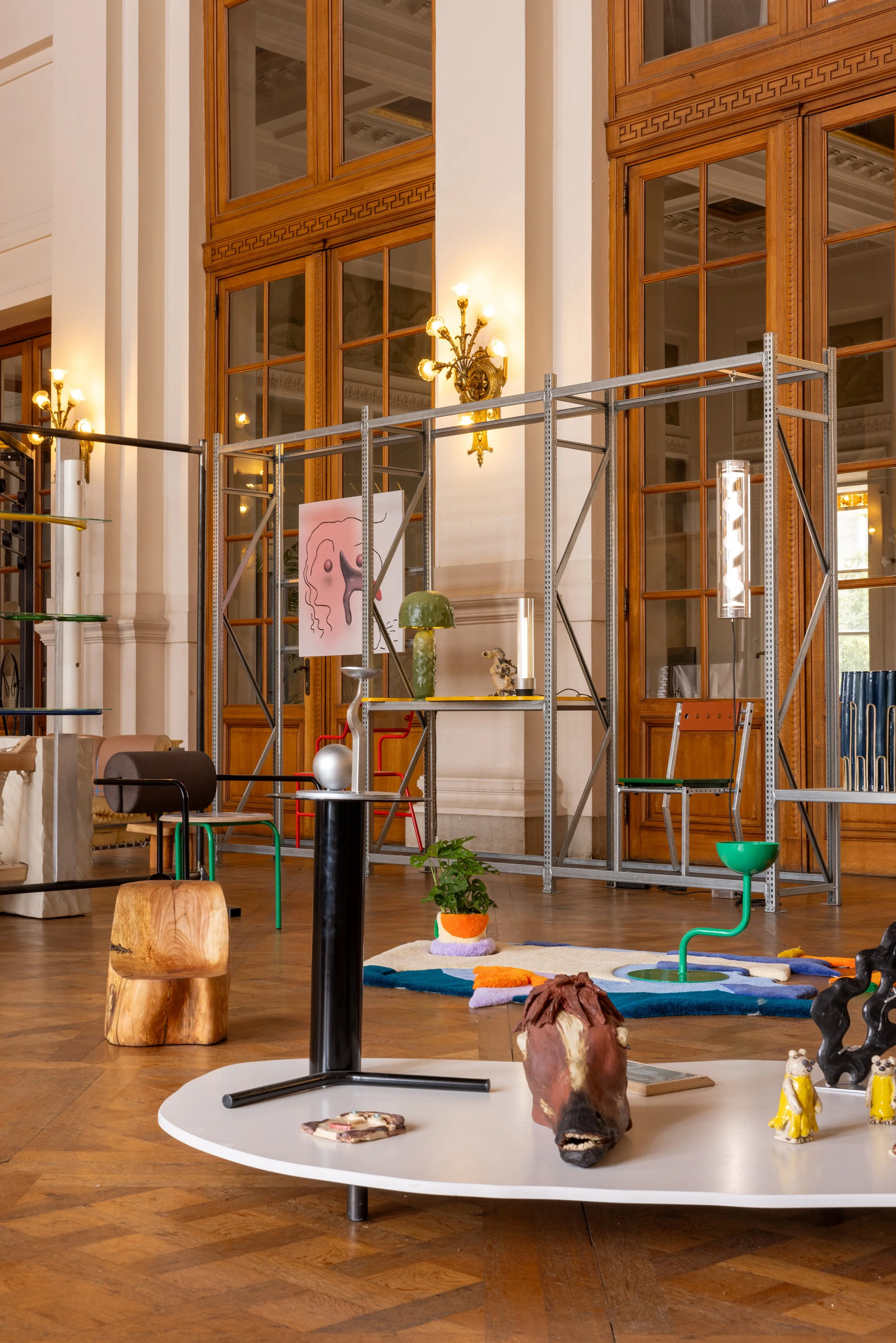 Interior of a building with wooden large window panels, ornate light fixtures, and various decorative objects and sculptures on tables and shelves. A scaffold is present indicating ongoing setup or renovation.