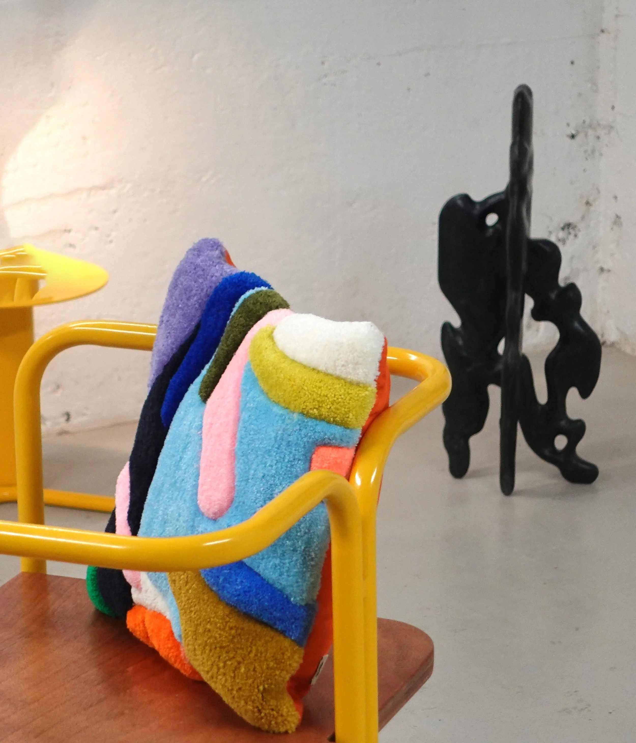 Colorful tufted cushion hanging on a yellow metal rack, with a black metal rack and a yellow plastic object on asurface nearby, against a white textured wall.