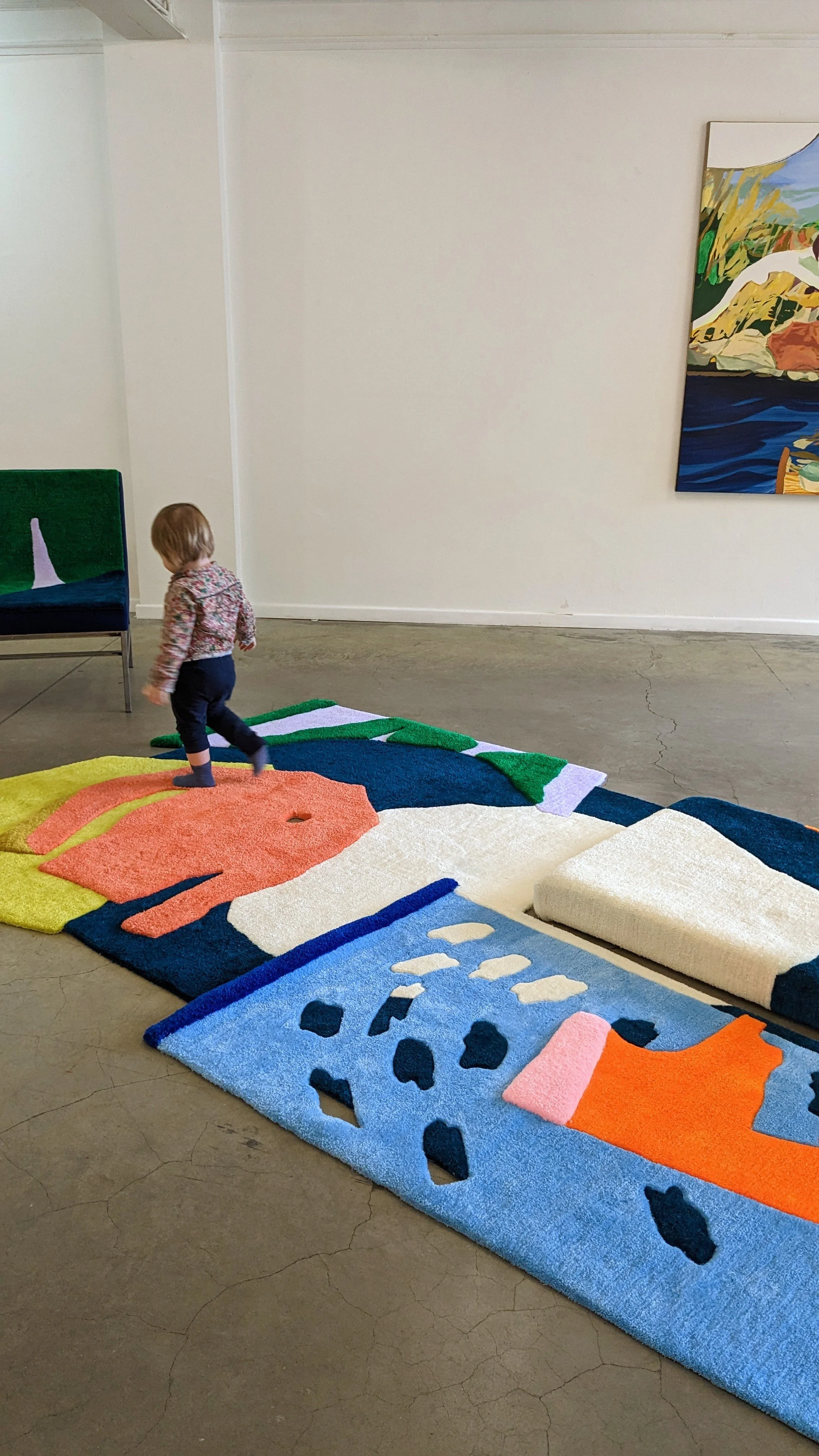 Child walking on colorful tufted abstract rugs in an art gallery with a painting hanging on the wall.