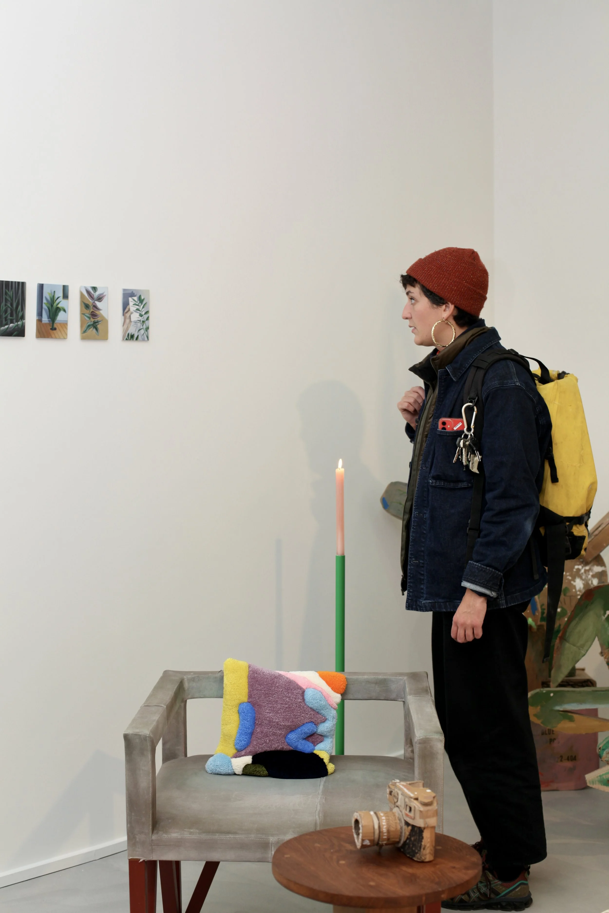 A woman wearing a red knit hat, hoop earrings, a dark jacket, and a backpack looks at artwork on a white gallery wall. The artwork consists of small, colorful paintings of plants. In the foreground, there is a concrete bench with a colorful textured 