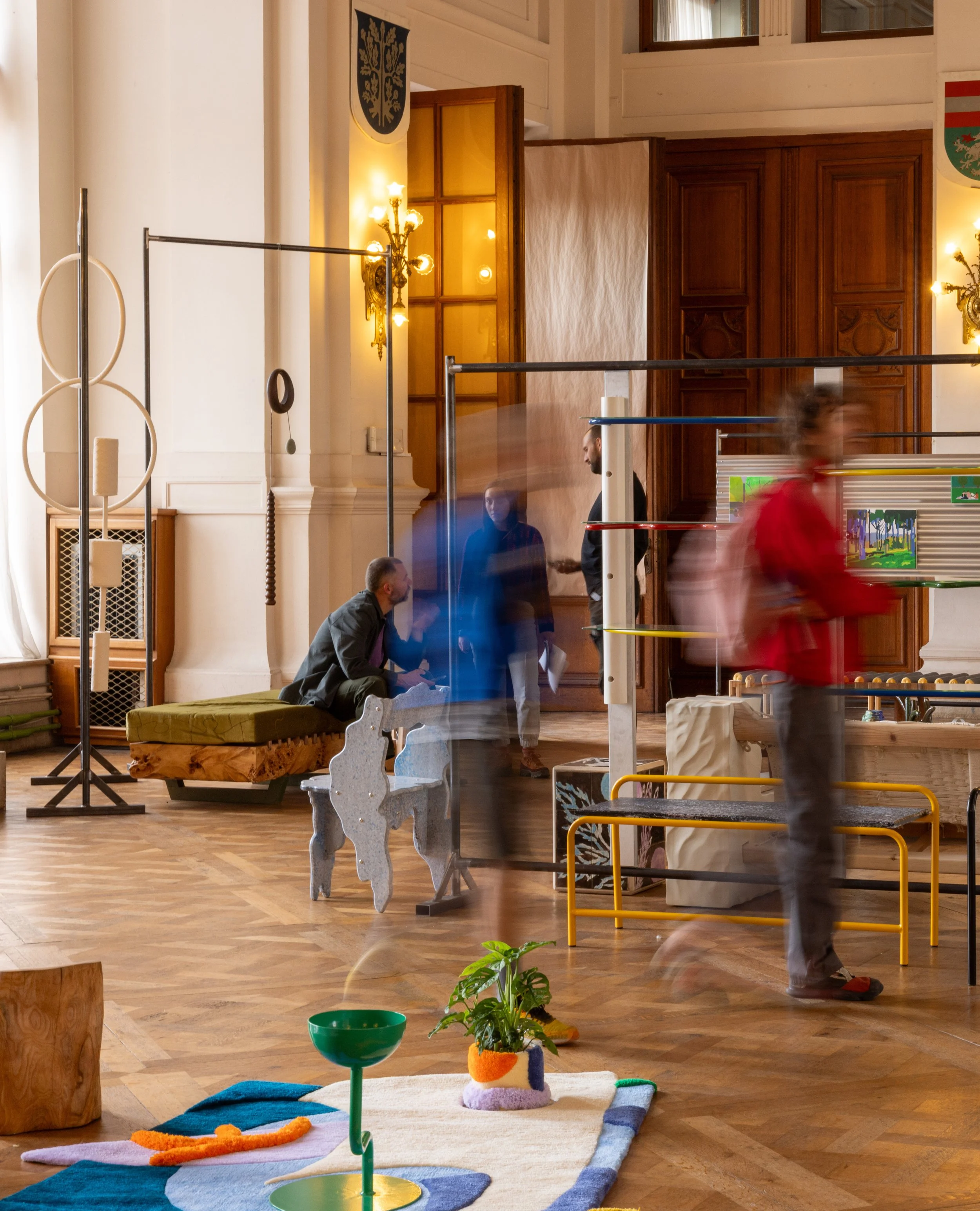 Indoor scene of an exhibition in La maison Communale de Saint-gilles, with colorful modern art and children playing with soft toys and a balancing table on a wooden floor.