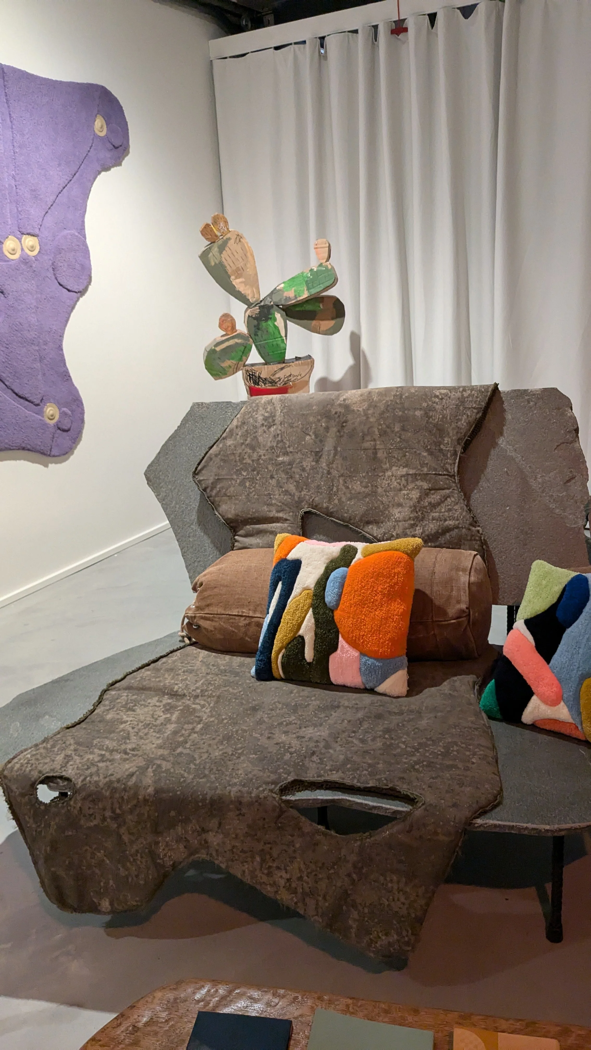 A cozy living room corner with a vintage brown armchair draped with a textured blanket. On the armchair are colorful, abstract tufted patterned pillows. Behind the chair is a cactus-shaped sculpture made of wood with painted green and tan sections. 