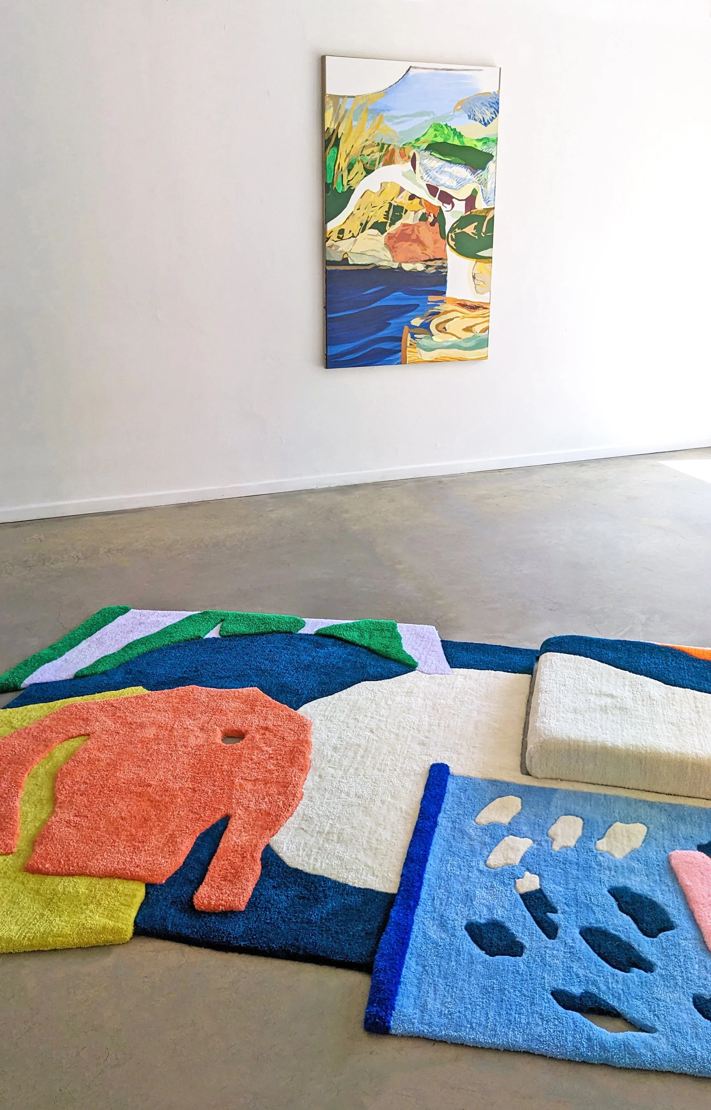 Colorful abstract painting on the wall with a white background and a heavily textured multicolored tufted rug on the floor featuring various bold shapes and patterns.