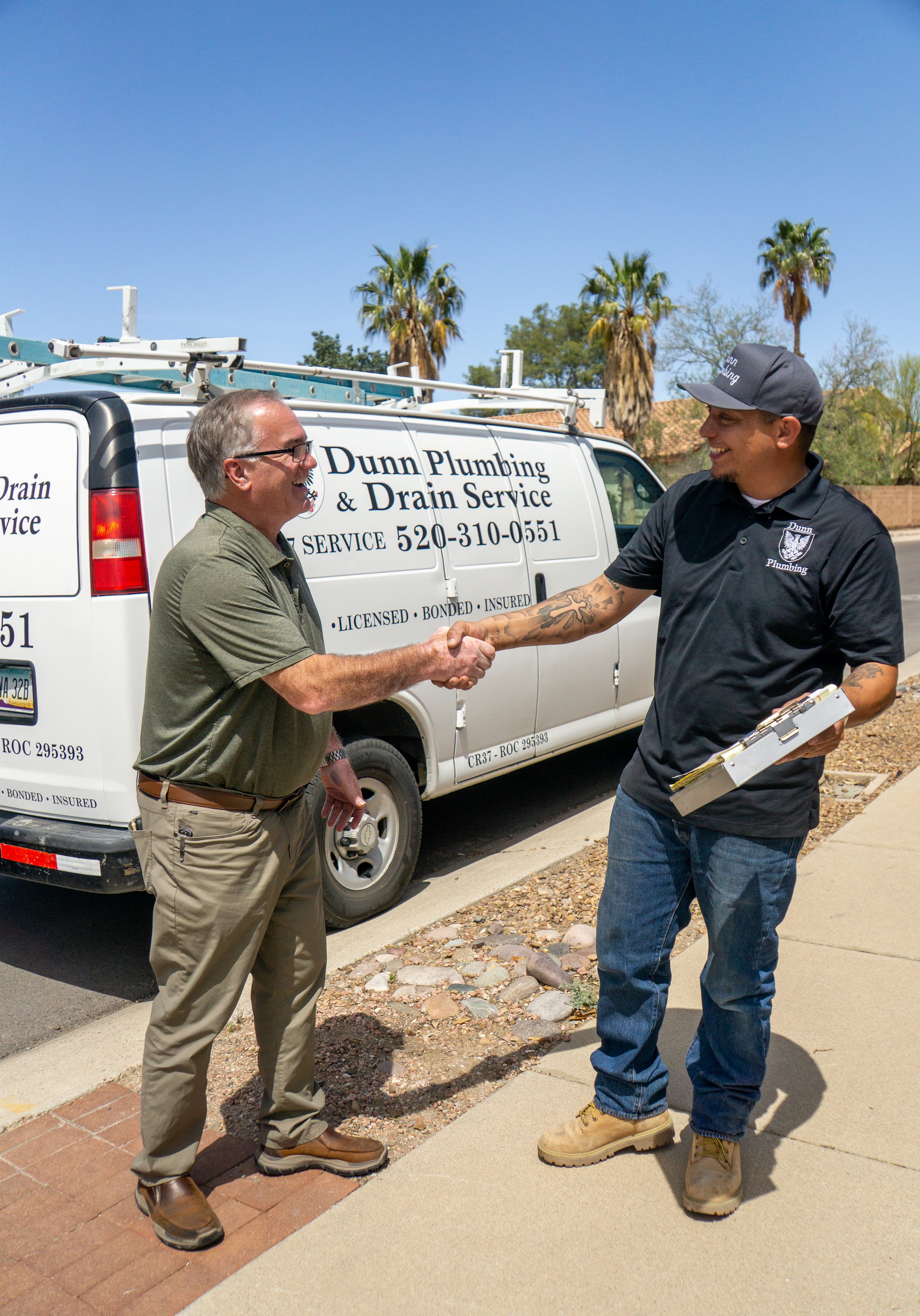 Two men shaking hands in front of a white service van labeled 'Dunn Plumbing & Drain Service,' with a clear blue sky and palm trees in the background.