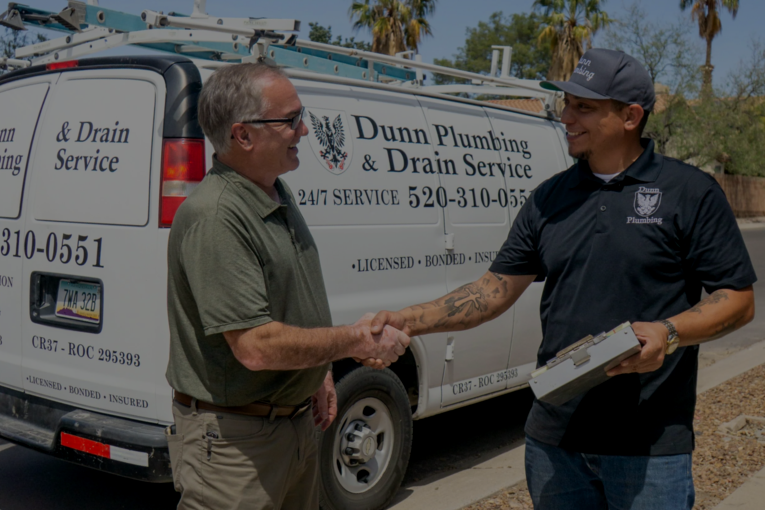 Two men shaking hands in front of a white work van with Dunn Plumbing & Drain Service logo, contact information, and services listed on it. One man is wearing glasses and a green polo shirt, the other man is in a black shirt and cap, holding a clipboard.