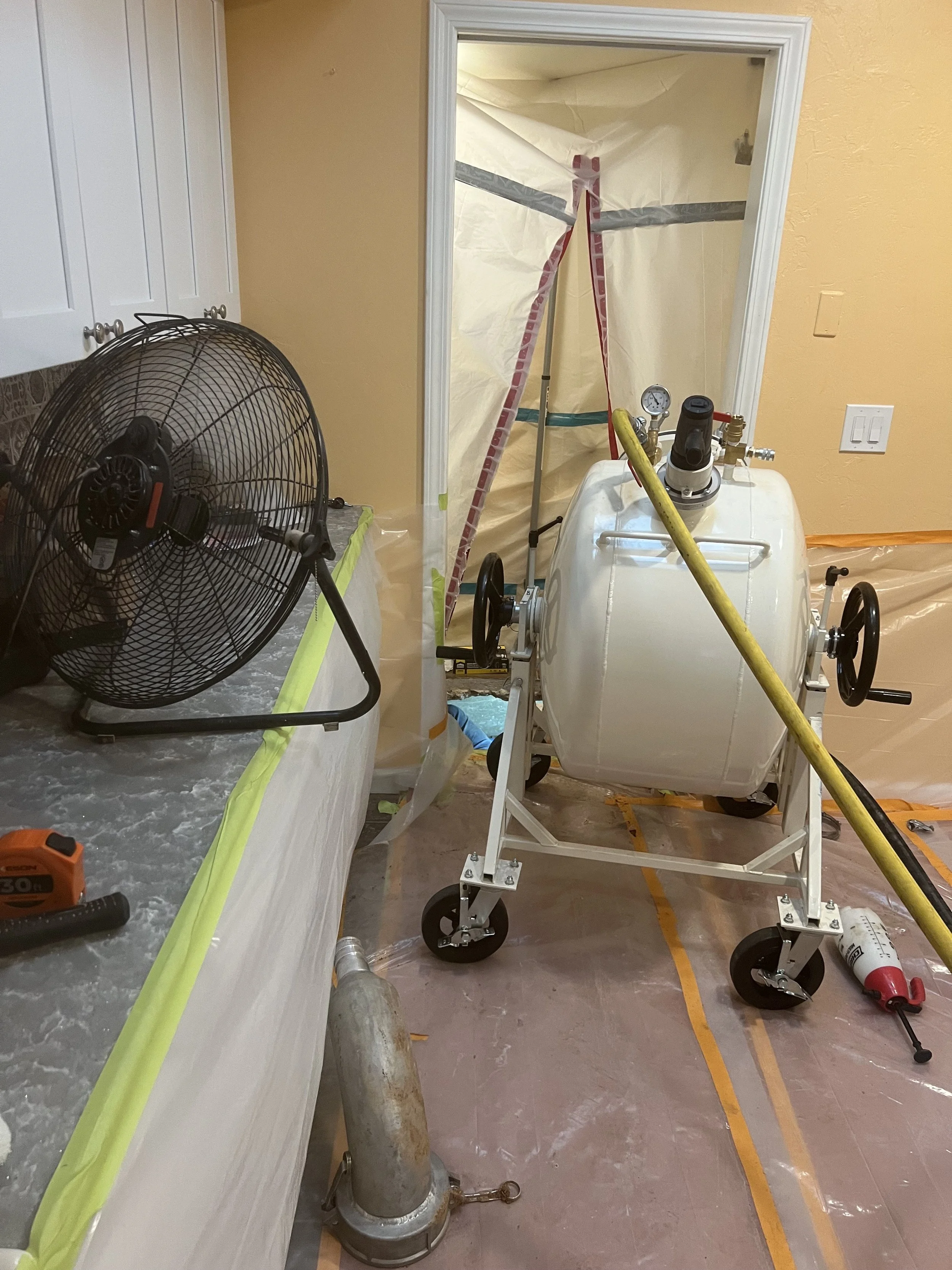 Construction workspace with a large white mixing machine on wheels, a black fan on a counter covered with a protective sheet, and orange tape on the floor marking off the area. Plastic sheeting covers the walls and doorway.