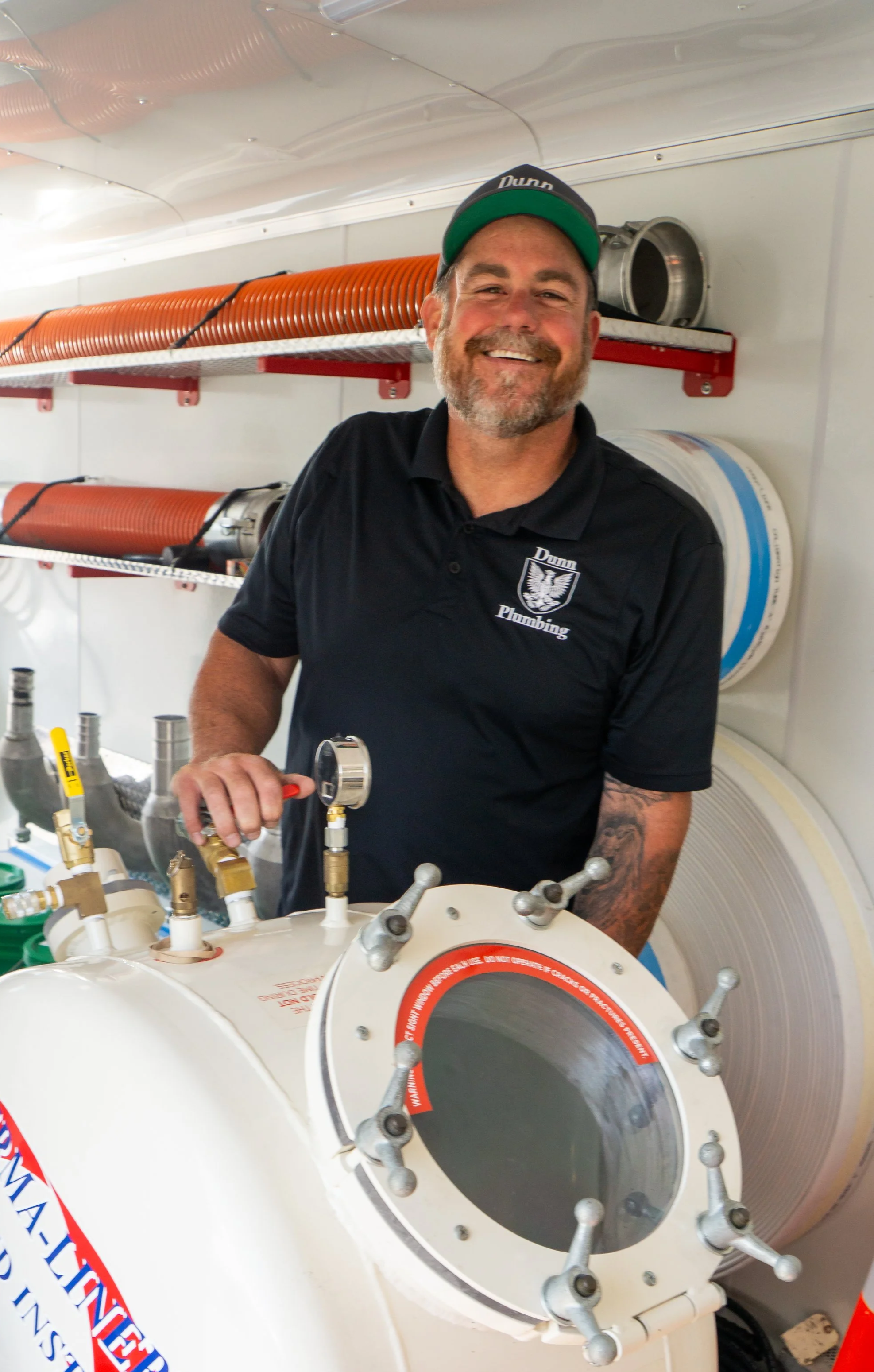 A smiling man in a black polo shirt and cap, operating a piece of plumbing equipment inside a maintenance truck or service van. The man is in front of orange and silver hoses mounted on the wall.