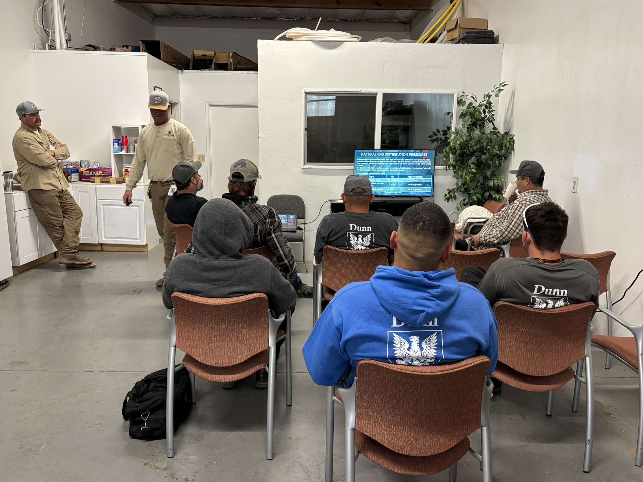 Group of people attending a presentation in a room with white walls, seated in chairs, watching a screen displaying information about natural gas distribution pressures. Two men stand near the back, one with arms crossed, the other walking.