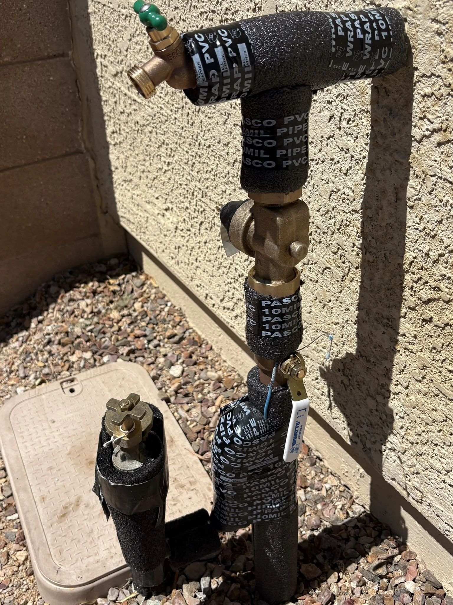 Outdoor home plumbing pipe with valve and pressure test tags, installed against a beige stucco wall, on gravel ground.