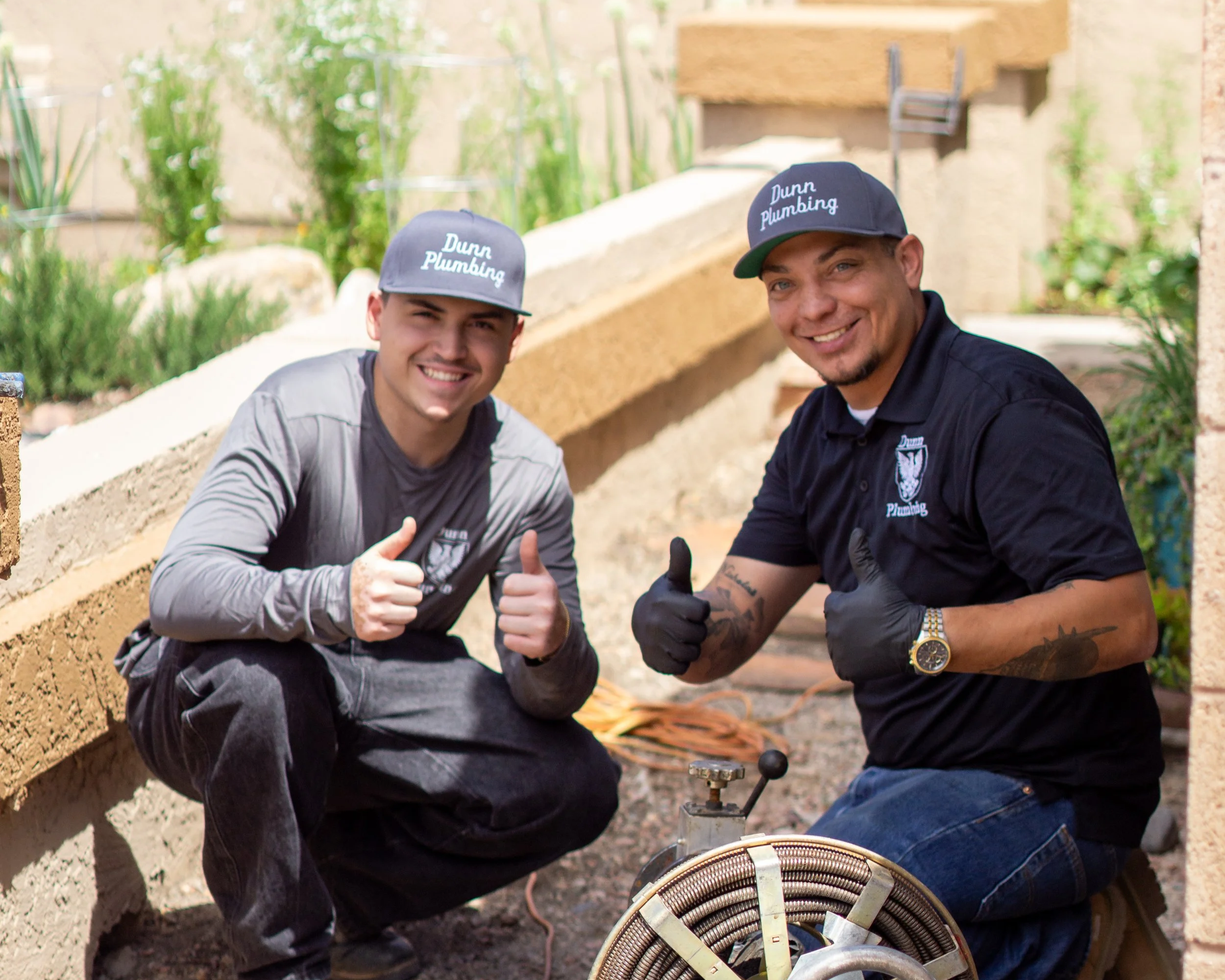 Two smiling professional plumbers in uniforms and caps squatting outdoors, giving thumbs-up gestures. They are surrounded by gardening plants and construction materials, with plumbing tools in front of them.