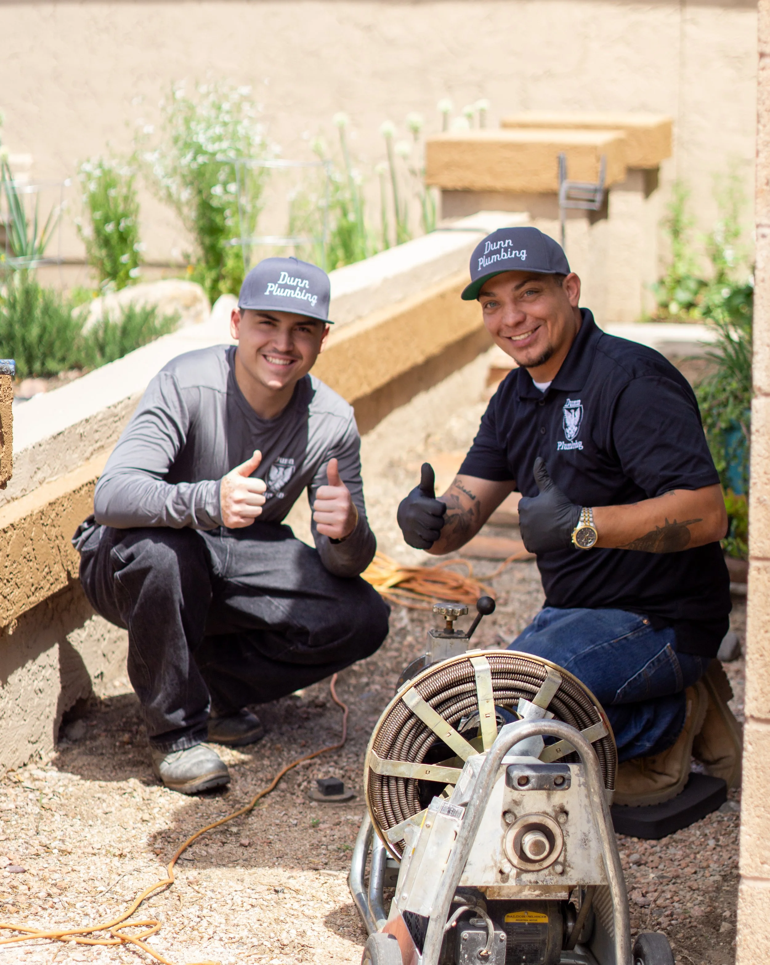 Two smiling men kneeling on the ground outdoors, giving thumbs-up, working on a construction or plumbing project, with a large coil of pipe in front of them, wearing caps and gloves.