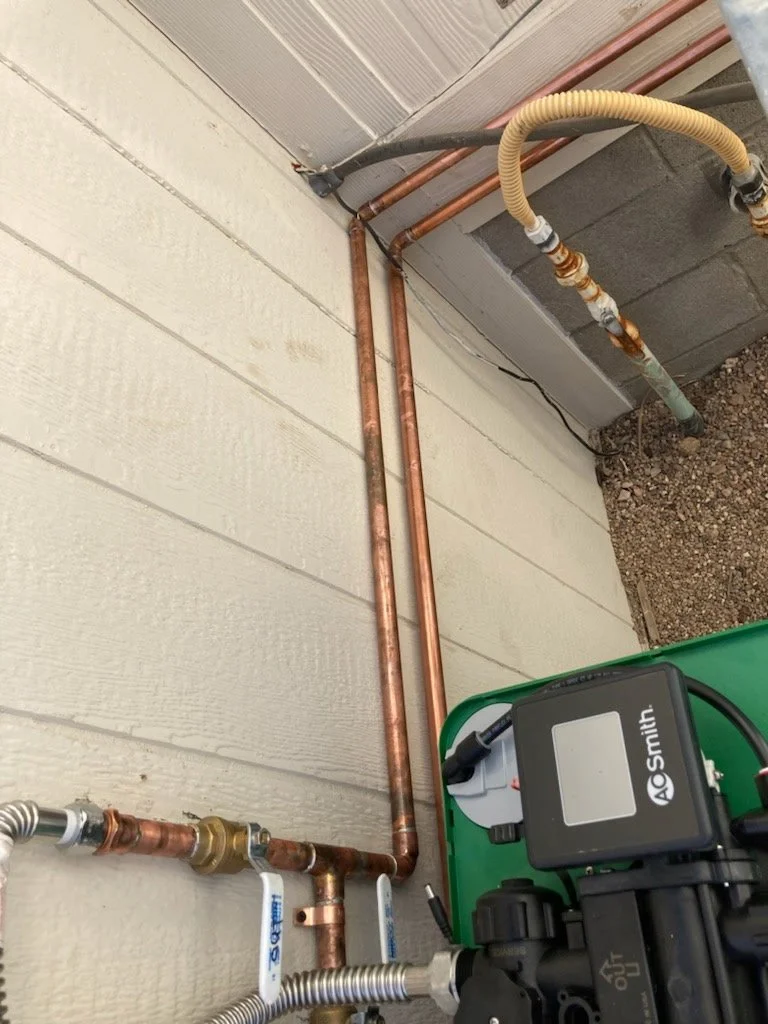 Close-up view of copper plumbing pipes and valves attached to the side of a house wall, with a green device and a control box nearby.