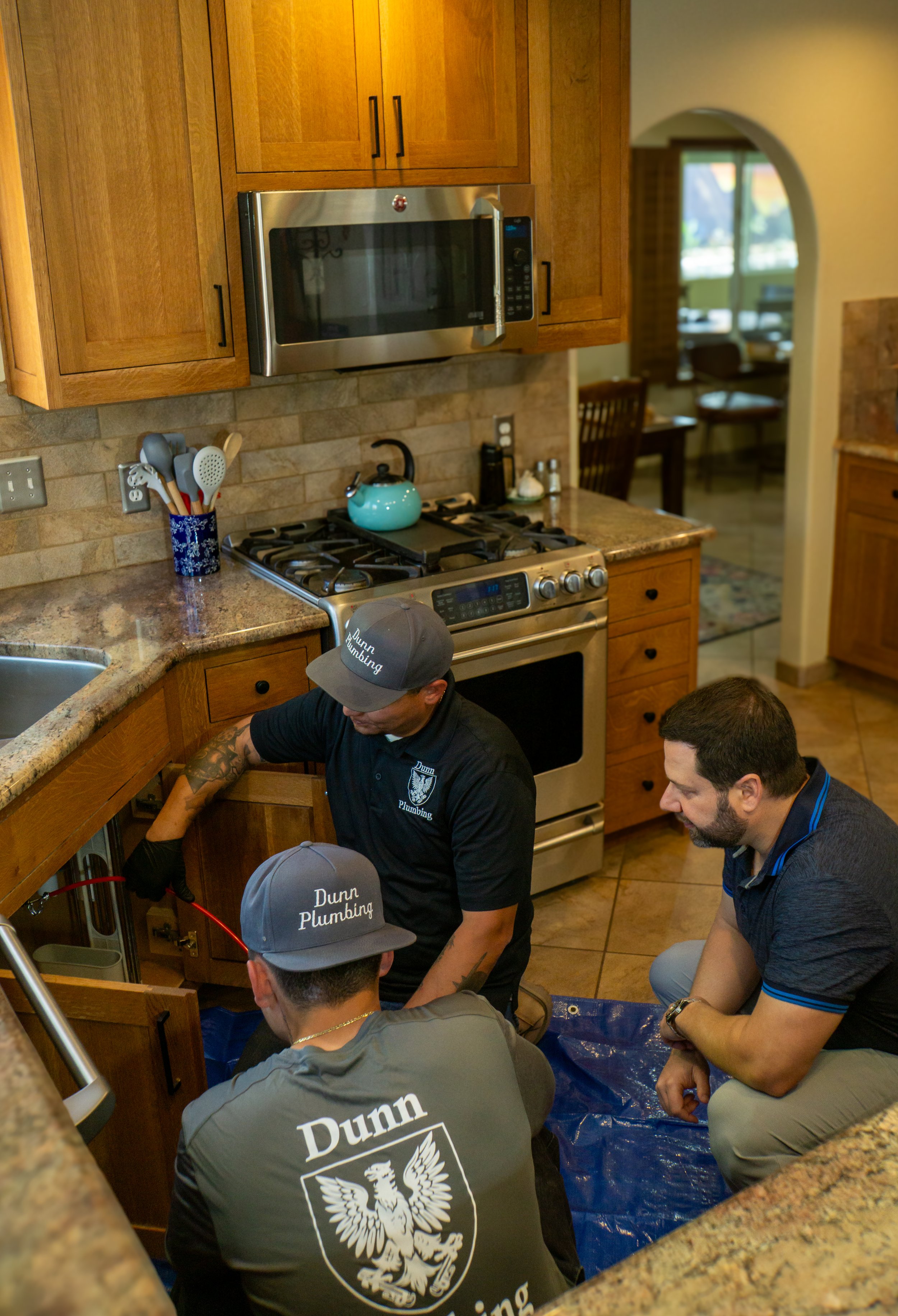 Three plumbers working under a kitchen sink, inspecting and repairing plumbing pipes. Two of them wear gray caps with 'Dunn Plumbing' logos and a gray vest with the same logo. The kitchen has wooden cabinets, a stove, and a microwave, with some utensils on the counter.