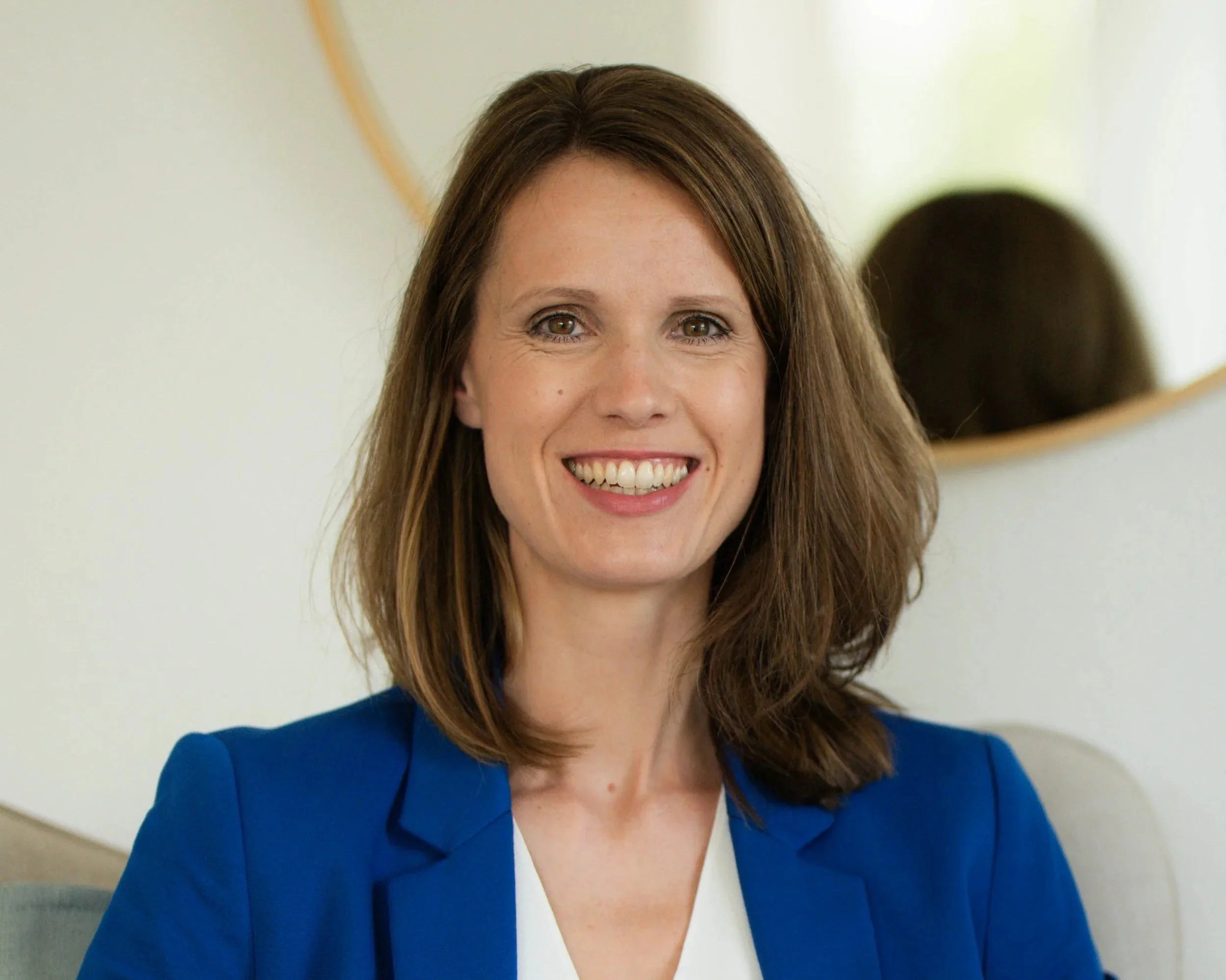Porträt einer lachenden Frau mit braunen Haaren, tragend blaue Jacke vor hellem Hintergrund.