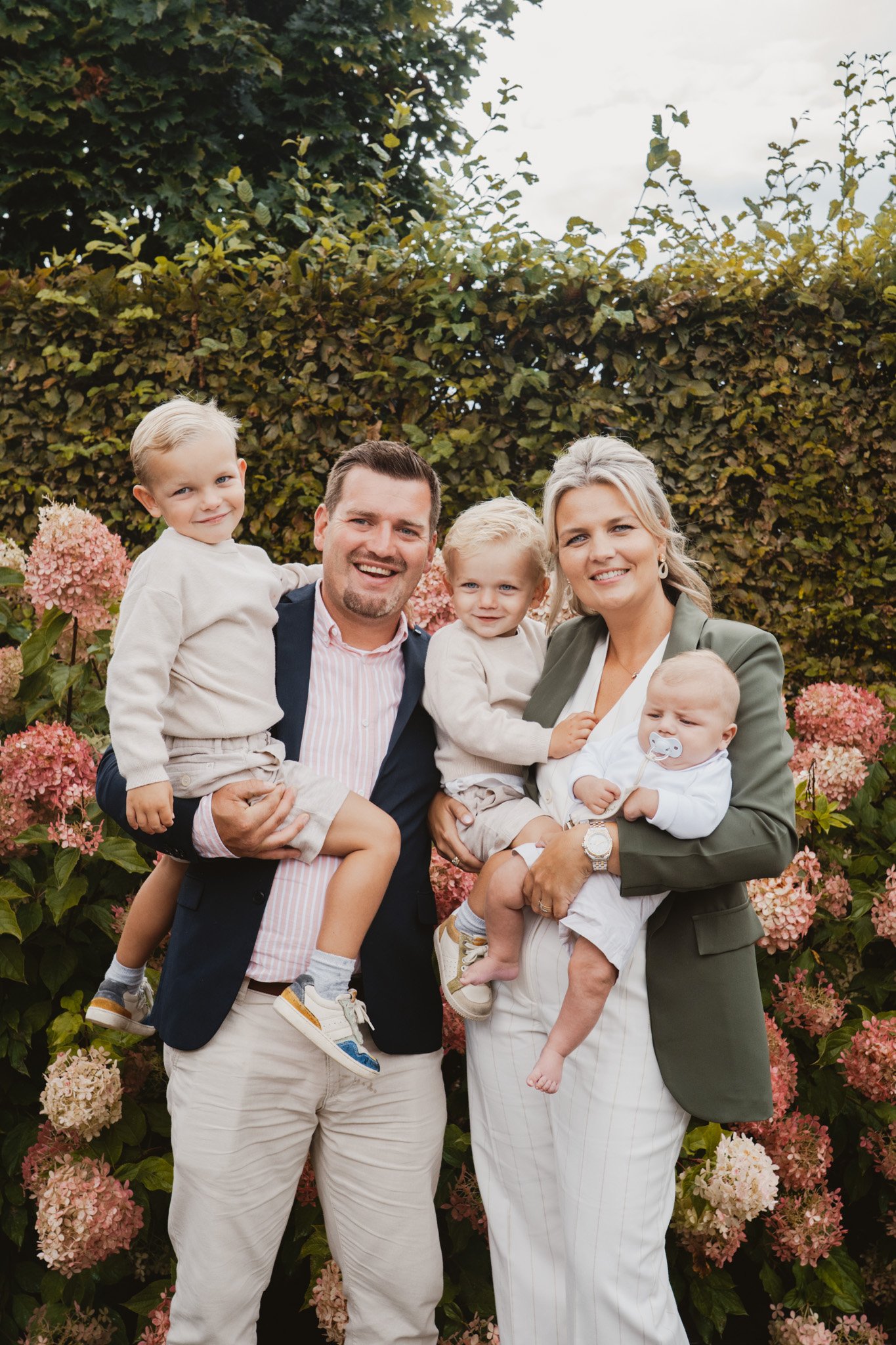 Gezin met vier kinderen poseert voor een bloemenhaag in een tuin, lachend en knuffelend.