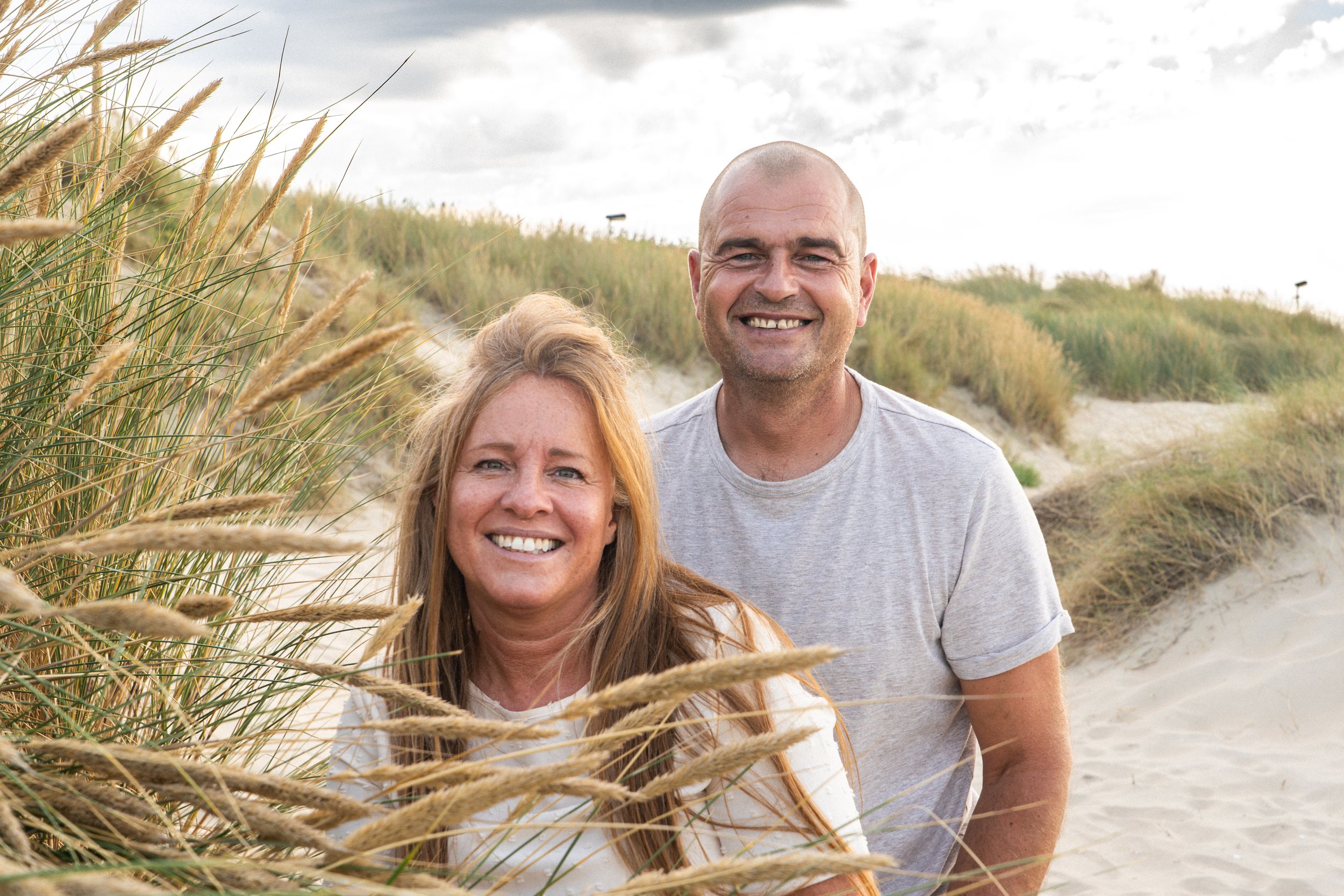 Een lachend paar op het strand, tussen grasvelden en zandduinen, onder een bewolkte hemel.