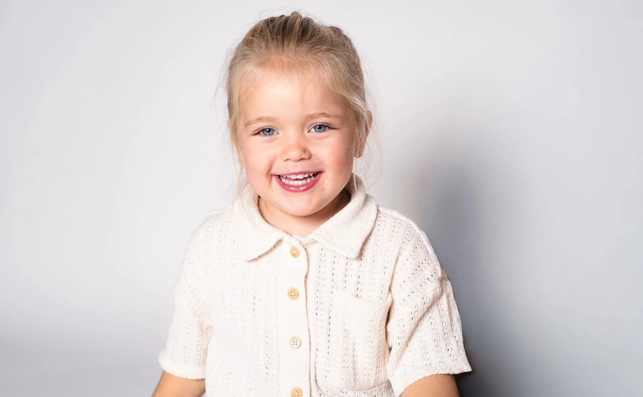 Een vrolijk lachend meisje met blonde haar en blauwe ogen, gekleed in een witte, gebreide blouse, staand voor een witte achtergrond.
