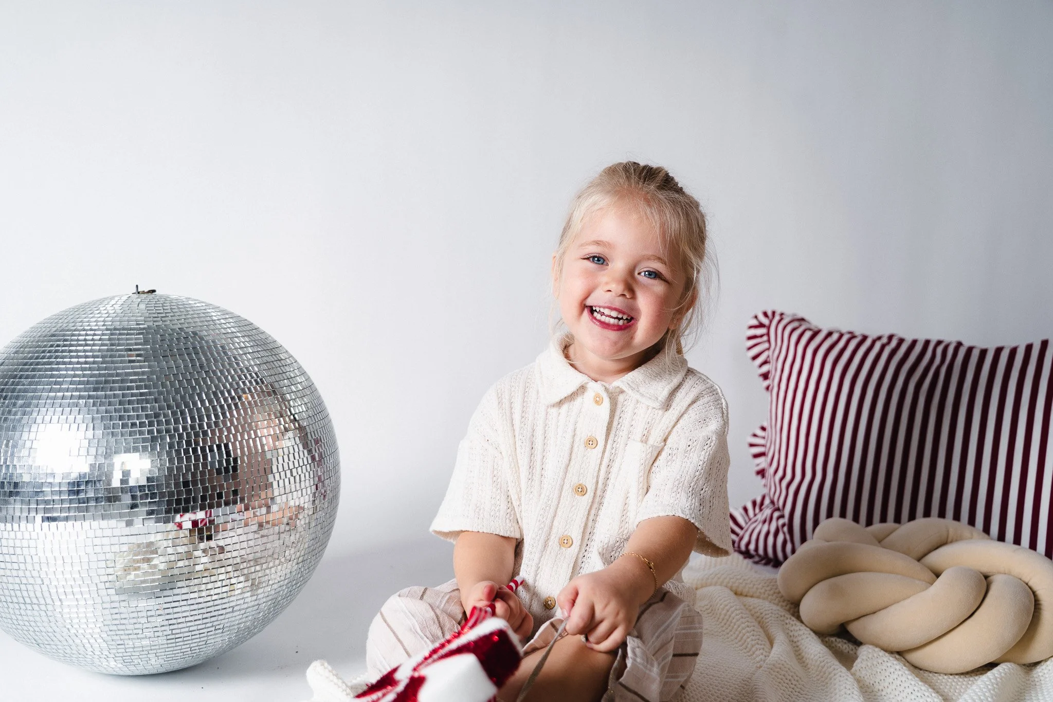 Lachende jonge meisje met blonde haar, zittend op een witdeken met een rode en witte knuffel, naast een grote spiegelbal en een rode en witte gestreepte kussen
